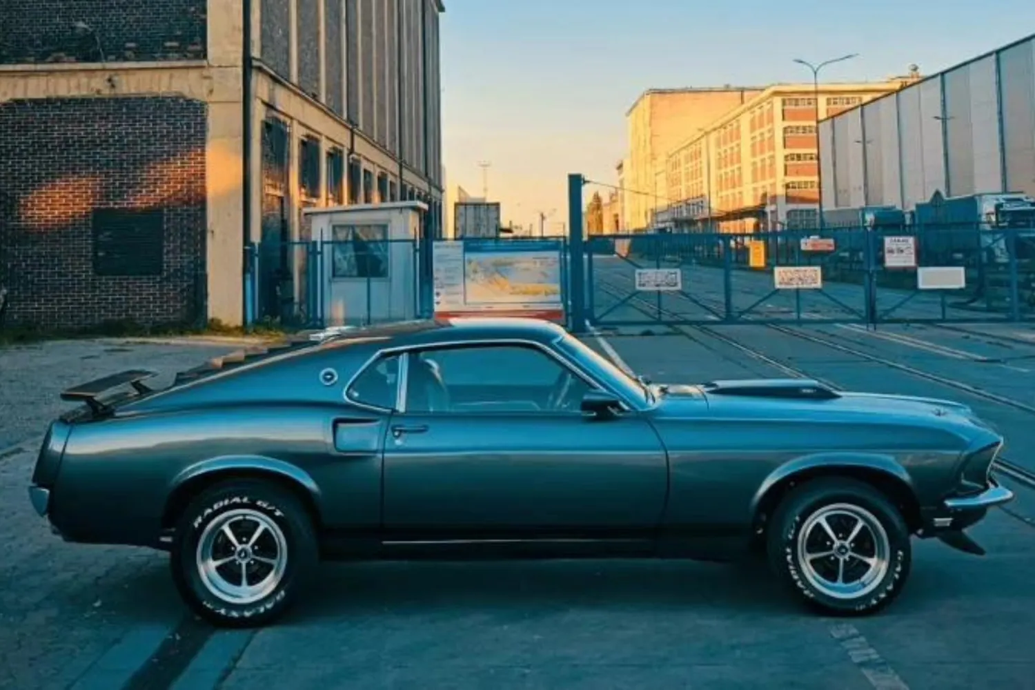 Grafitowy Ford Mustang Mach 1 z 1969 roku widziany z boku, przed bramą i ceglanymi halami