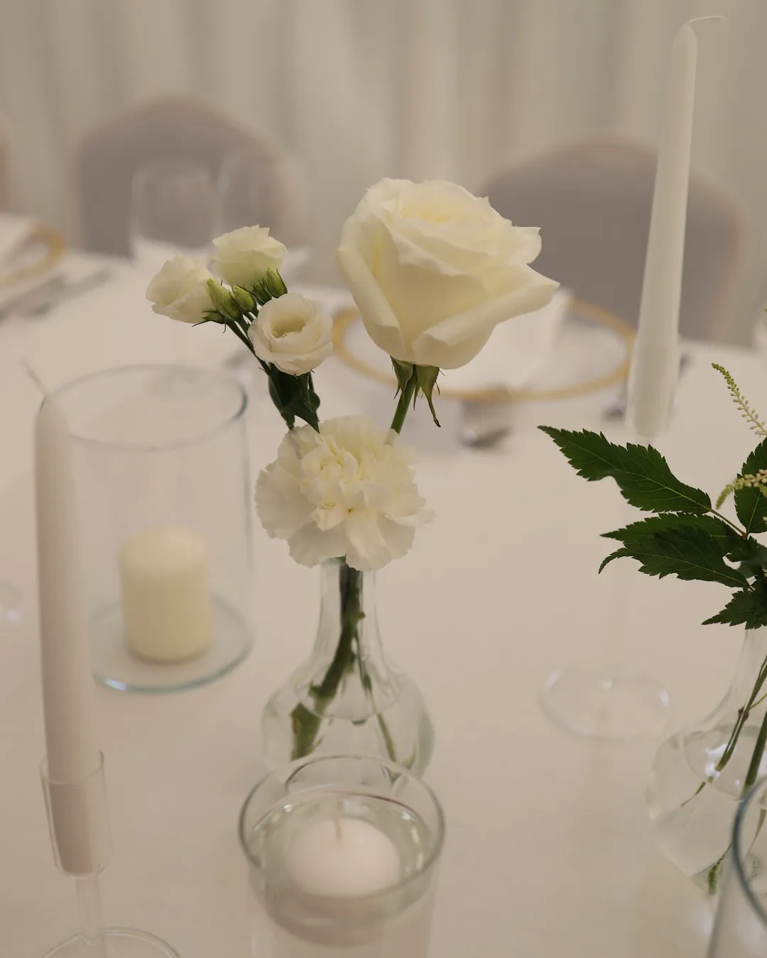 White rose, eustoma and carnation in a glass vase next to candles in glass and a floating candle on a white tablecloth