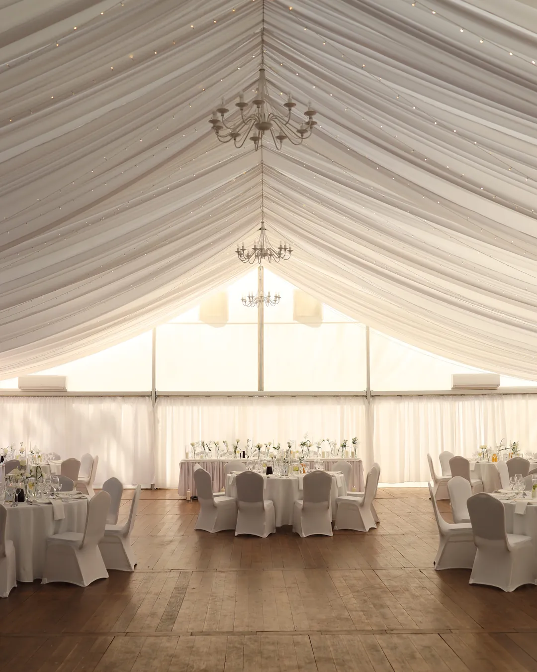 The interior of a white pavilion with a fabric ceiling, chandeliers and small lamps above round tables