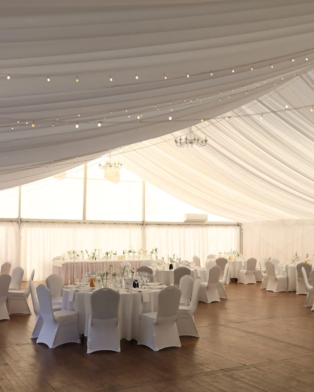 Strings of small lamps woven into the white fabric under the pavilion ceiling, above the laid tables and chandeliers