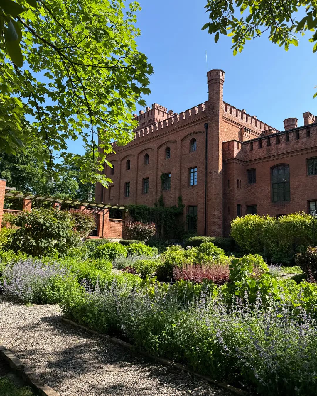 Brick castle behind garden beds with purple flowers, gravel walkway and pergola with hanging lanterns