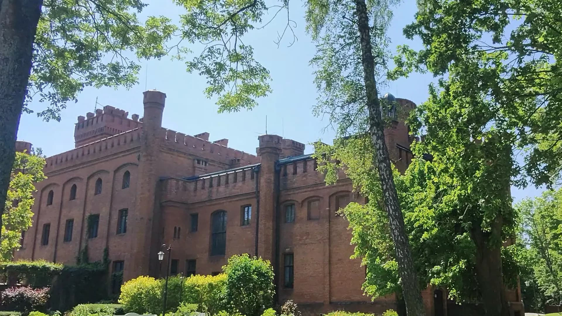 A red brick castle among the tall trees of the park, in front of it garden beds and green bushes