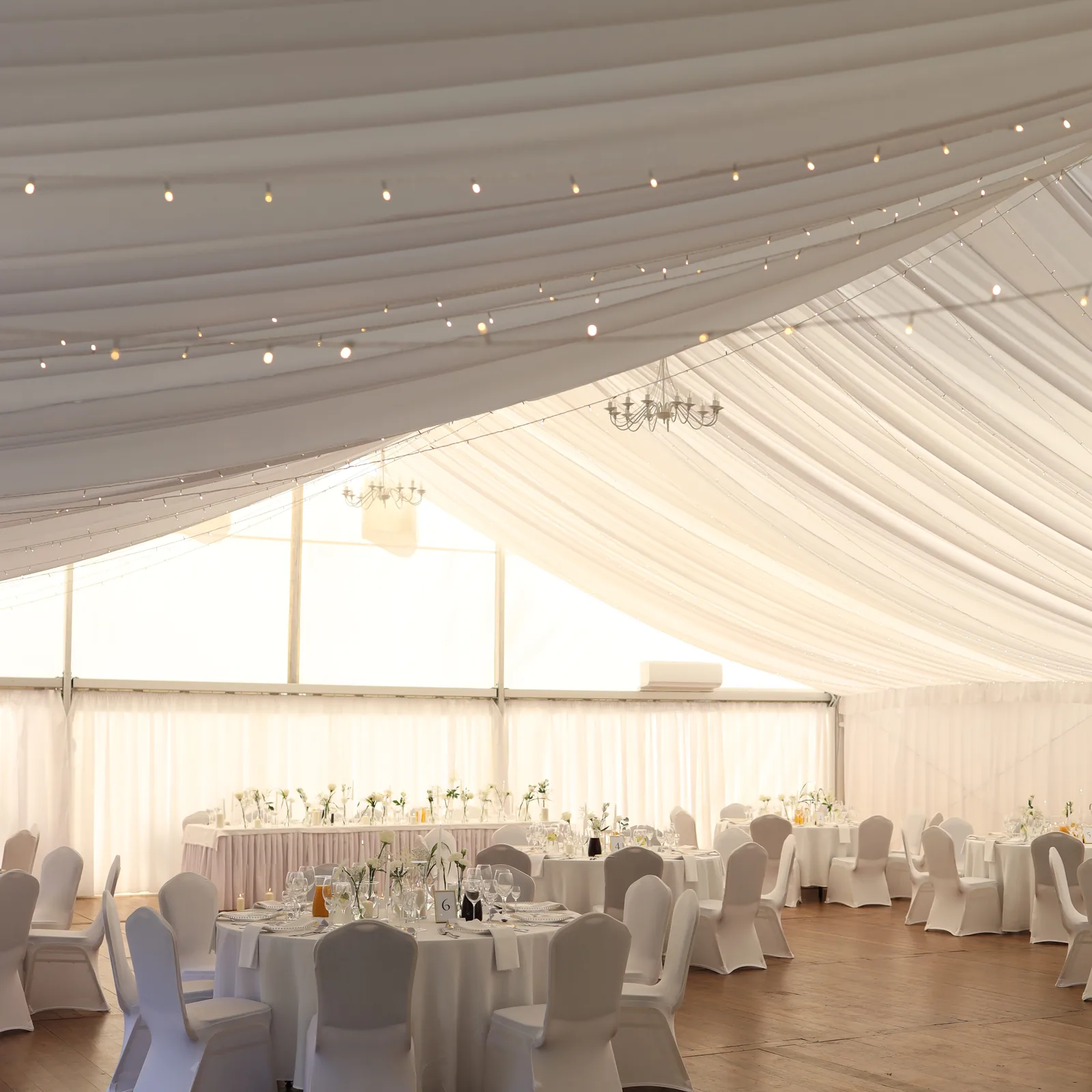 Garden pavilion on the wedding day: tables with white tablecloths, chairs with white covers, flowers in glass and strings of lights woven into the fabric under the ceiling