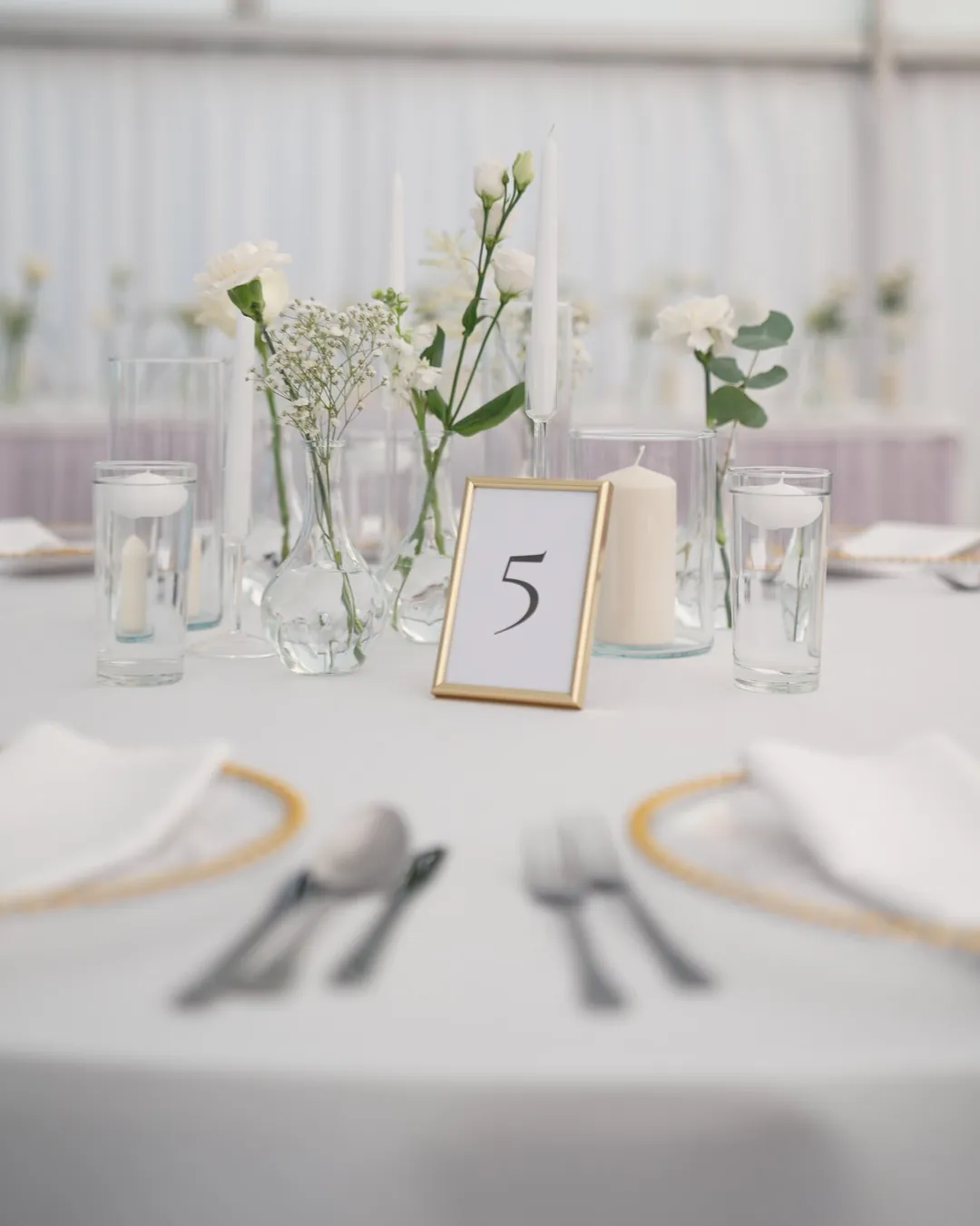 Round table with white tablecloth, glass charger plates, flowers in vases and table number in a gold frame