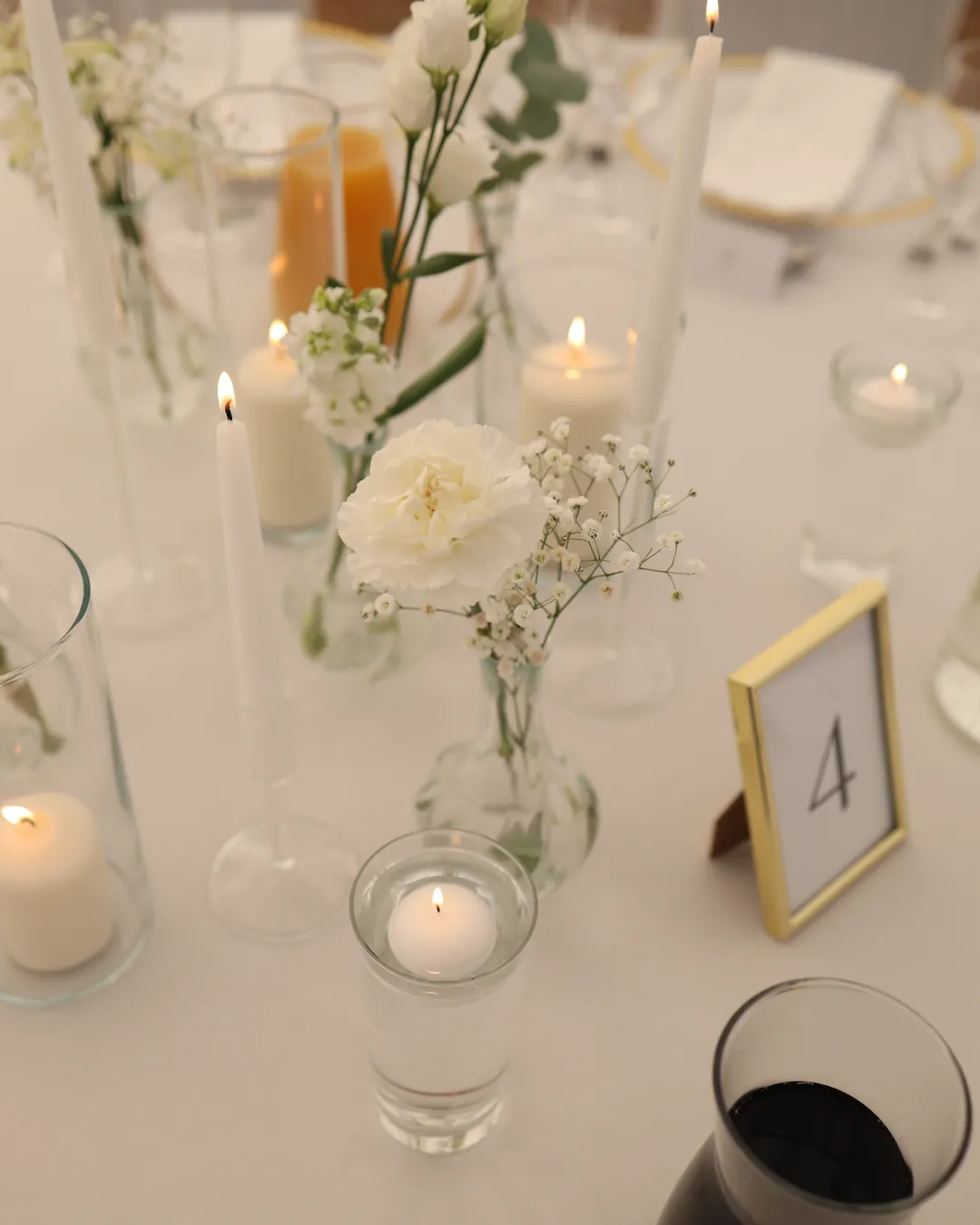 Lit candles in glass cylinders and a floating candle, next to it there is a carnation with gypsophila and a table number in a gold frame