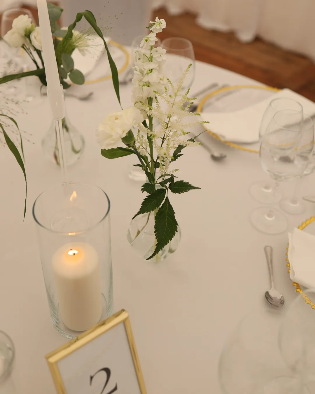 A candle in a glass cylinder, a vase with white flowers and astilbe and a gold frame with a table number on the tablecloth
