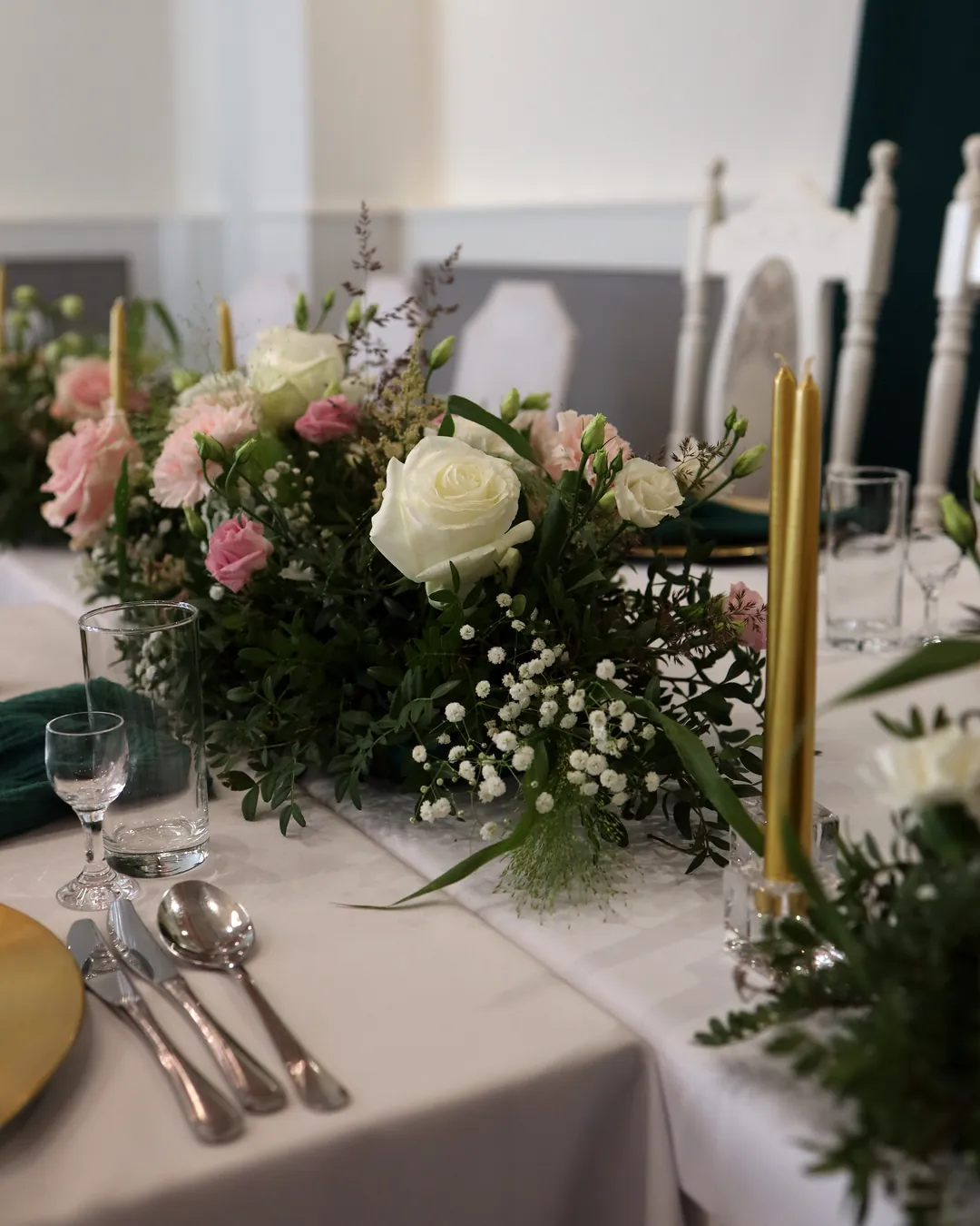 A low composition with a white rose, pink carnations and gypsophila on the front of the couple's table, next to it are thin gold candles in glass candlesticks
