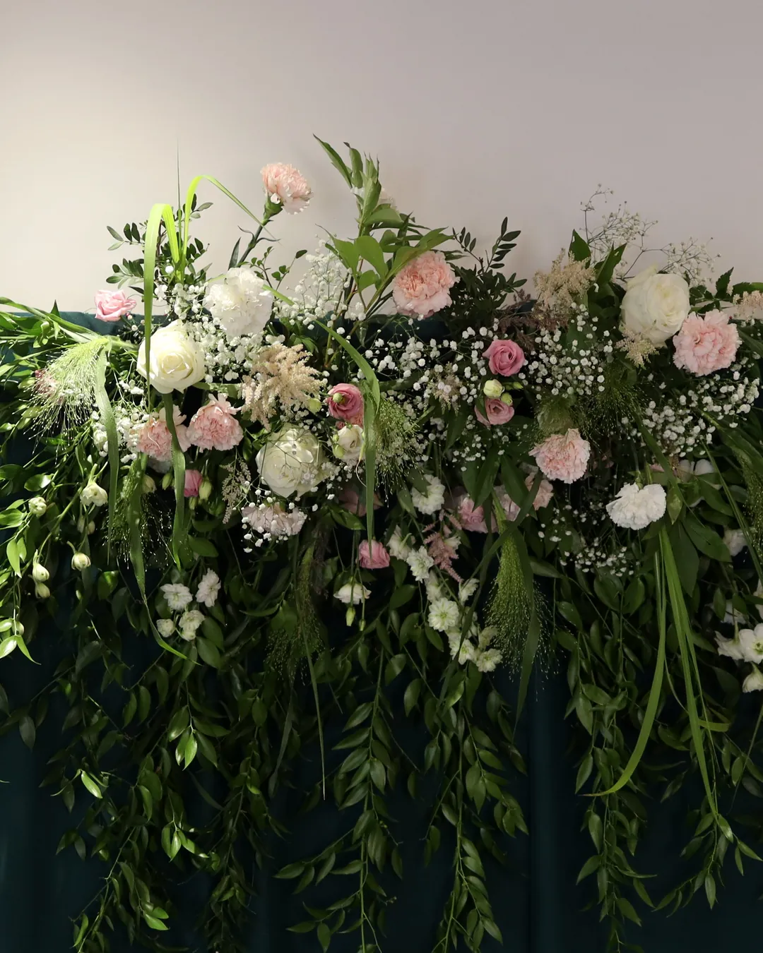 A garland of roses, carnations, gypsophila and grasses at the top of the bottle-green wall, with ruscus shoots flowing down the fabric