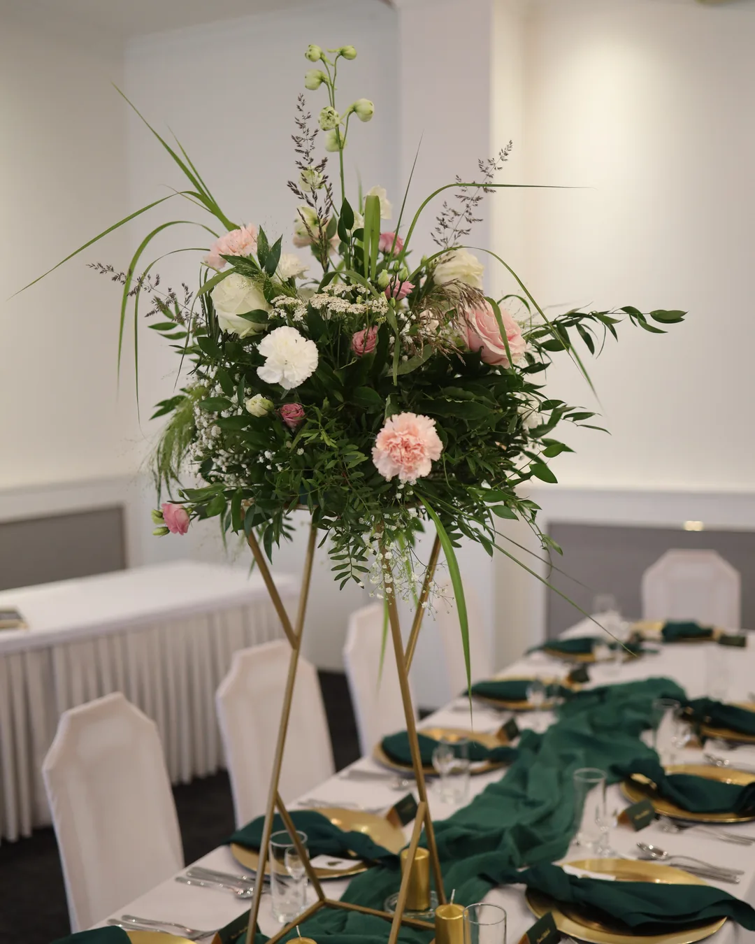 Tall composition on a golden geometric stand: roses, carnations, gypsophila and grasses over a table with a bottle-green table runner