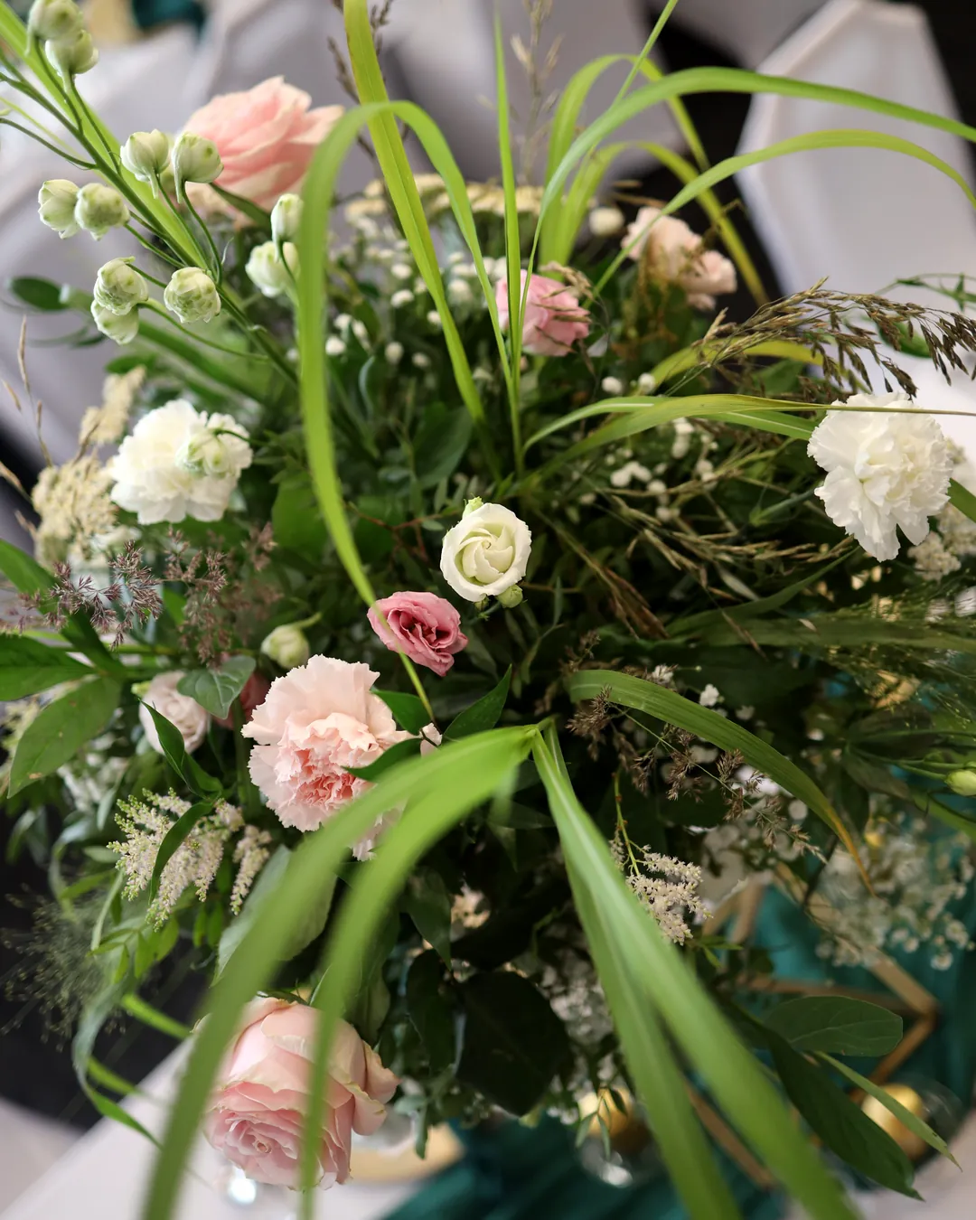 Light pink roses, white carnations, eustoma and long grasses in a tall composition above the guests' table with a green table runner