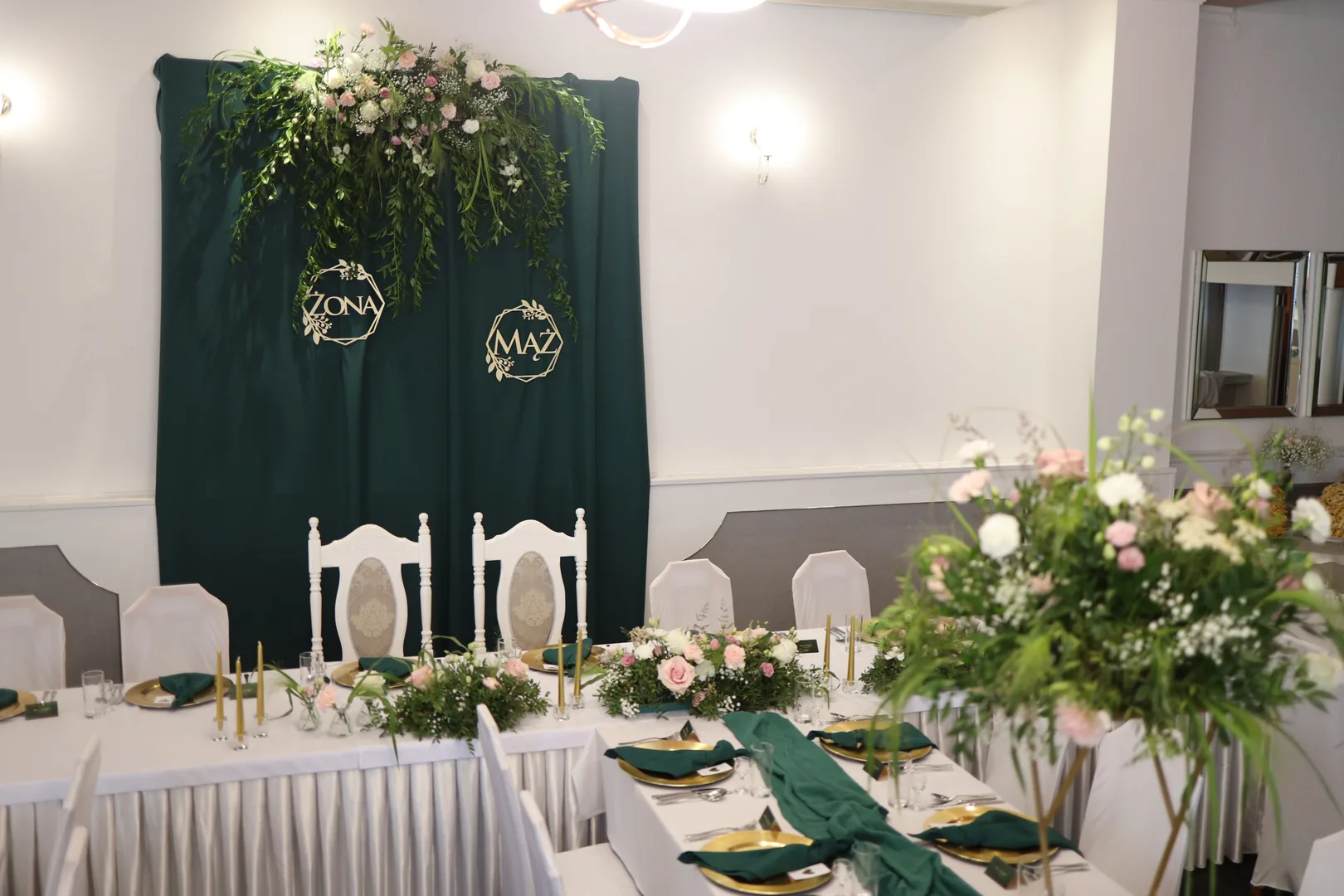 The bride and groom's table with white carved chairs, low compositions and golden candles, behind it a green wall with a garland