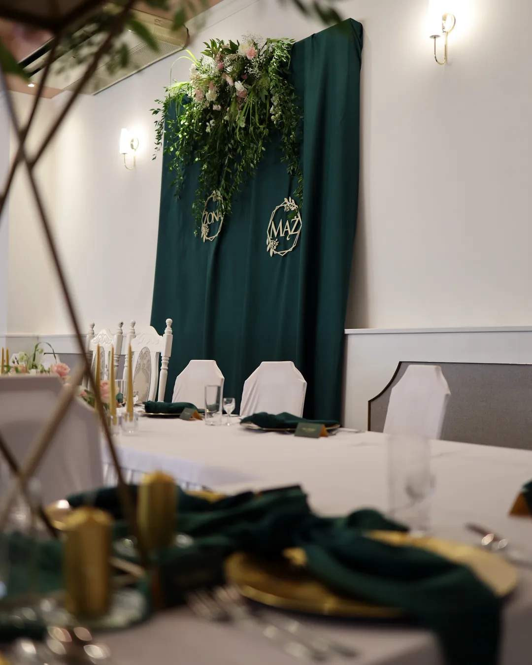 A green wall with a garland and gold hoops seen from the guest's seat, gold candles, plates and napkins in the foreground