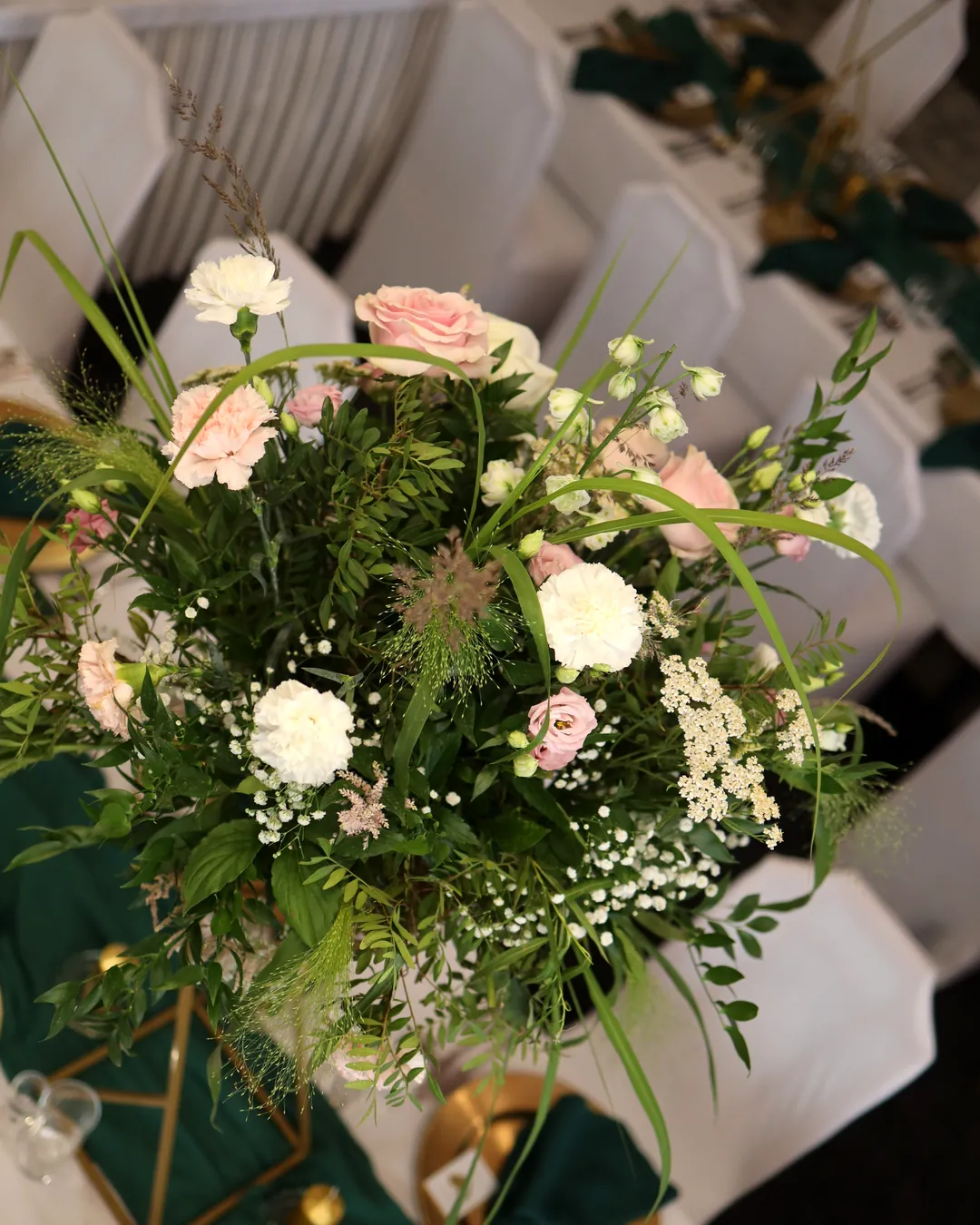 A tall arrangement of light pink roses, white carnations, gypsophila and grasses seen from above, above a gold stand and green table runner