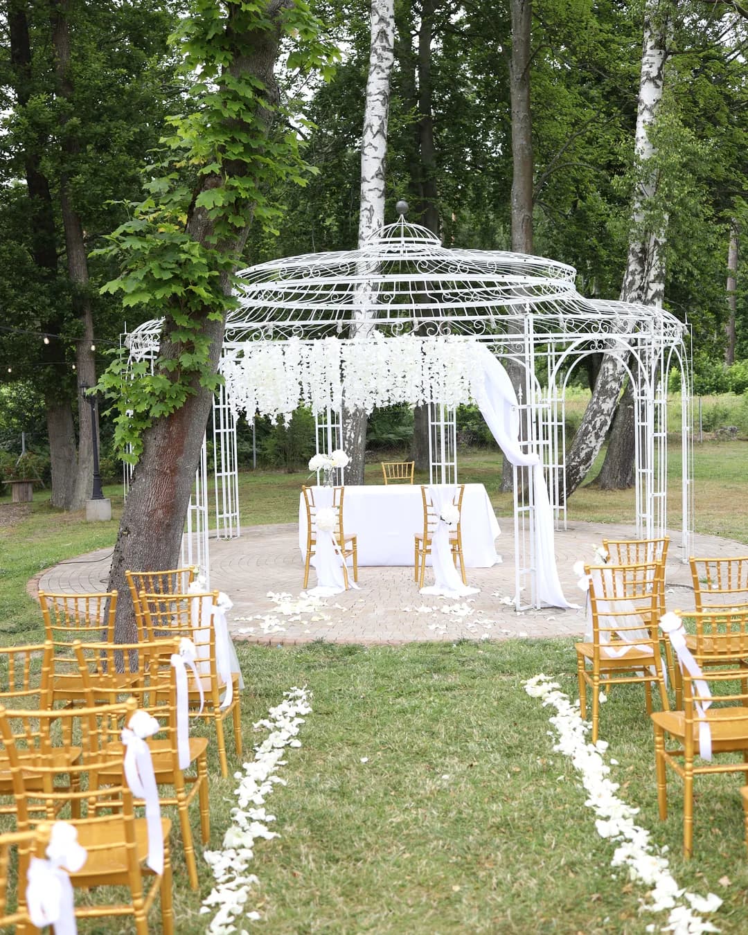 Outdoor ceremony: an alley strewn with rose petals, gold chairs with tulle bows and a white gazebo with a table
