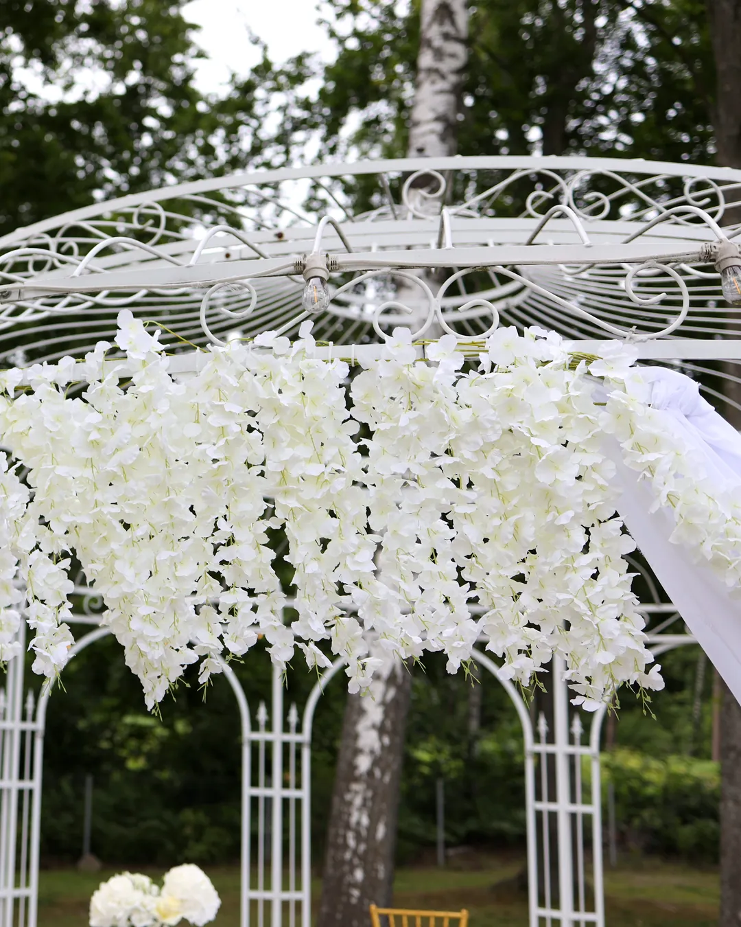 White openwork gazebo in the park with a garland of white flowers, light bulbs and flowing white fabric against the background of birches