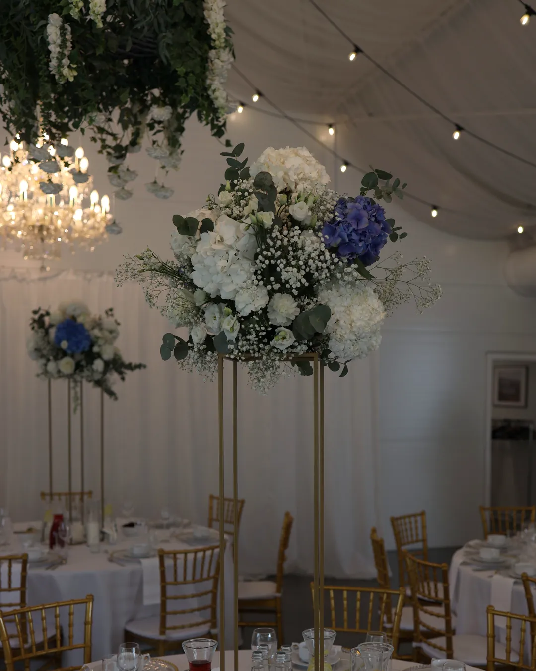 A tall golden flower stand with white and blue hydrangeas under a fabric with light bulbs, next to it a crystal chandelier and a green wreath