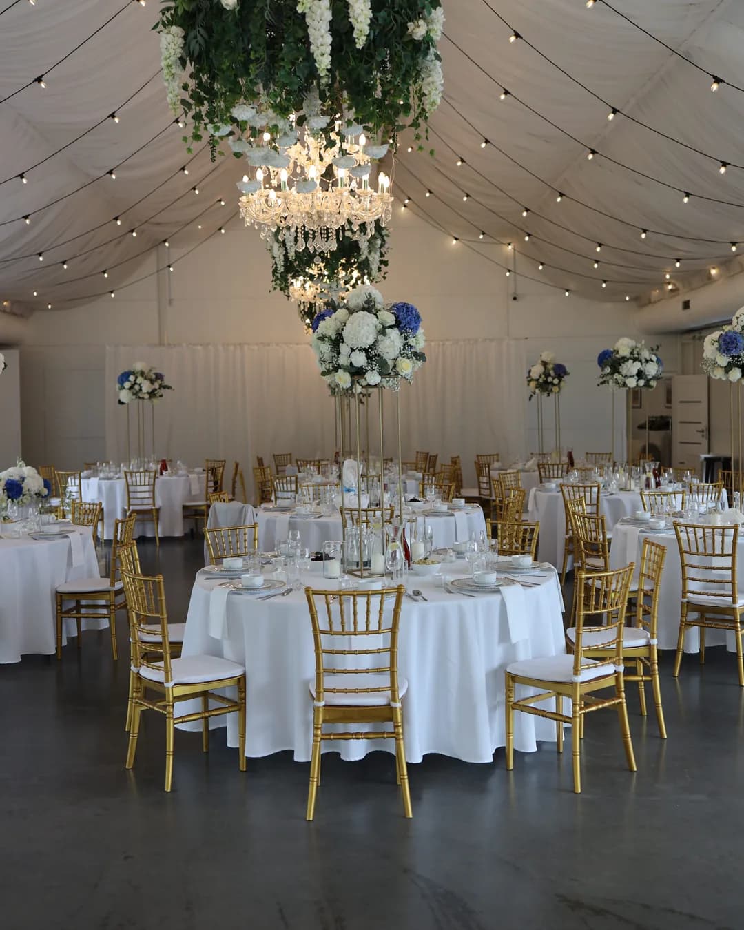Wedding hall with round tables covered with white tablecloths and tall golden flower stands with white and blue arrangements