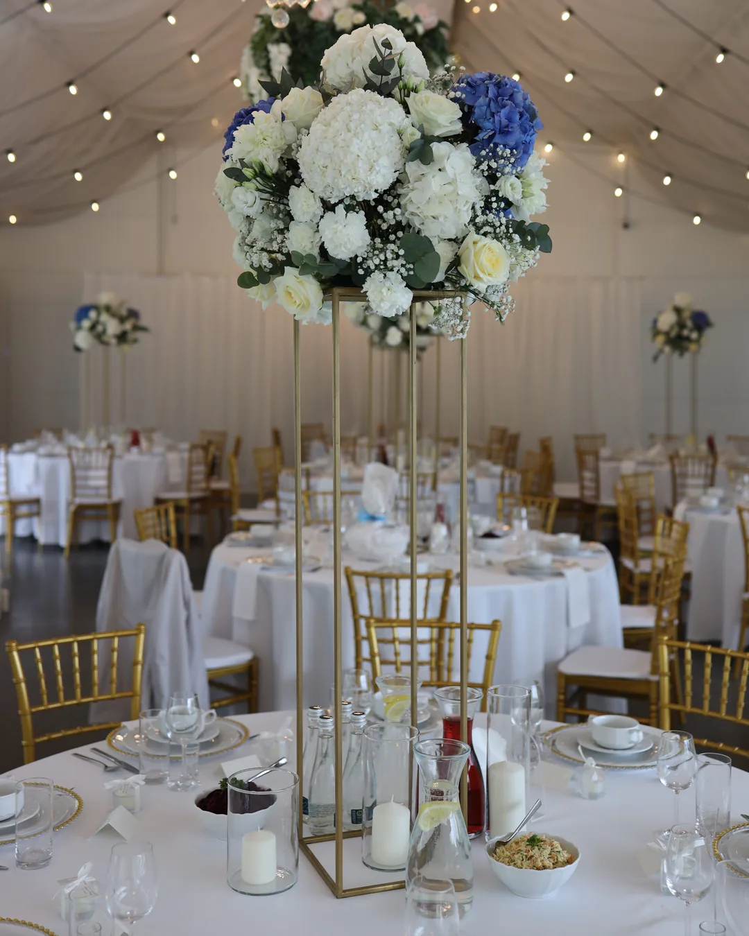 Guest table with a composition of hydrangeas, roses, carnations and gypsophila on a golden flower stand and pillar candles in glass
