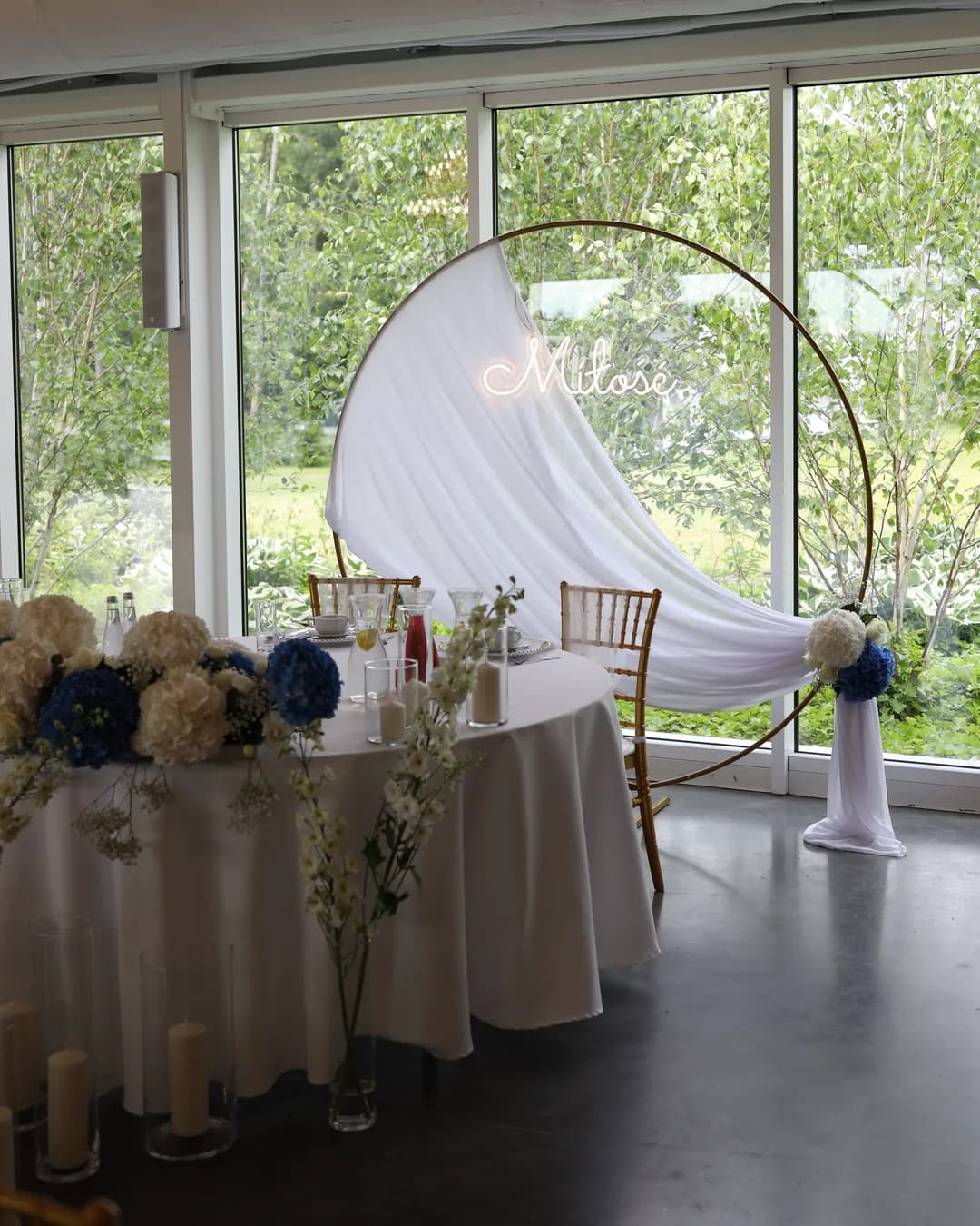 The bride and groom's table from the side: a composition of hydrangeas on the front, vases with white delphinium and candles in glass near the glass wall