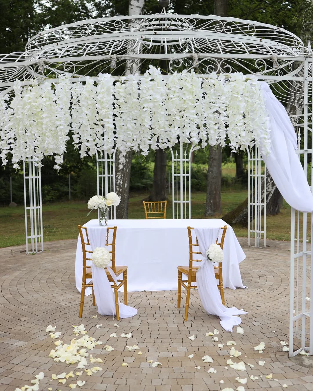 Ceremony table under a white arbor, couple's chairs with white chiffon and hydrangea, rose petals on a stone circle