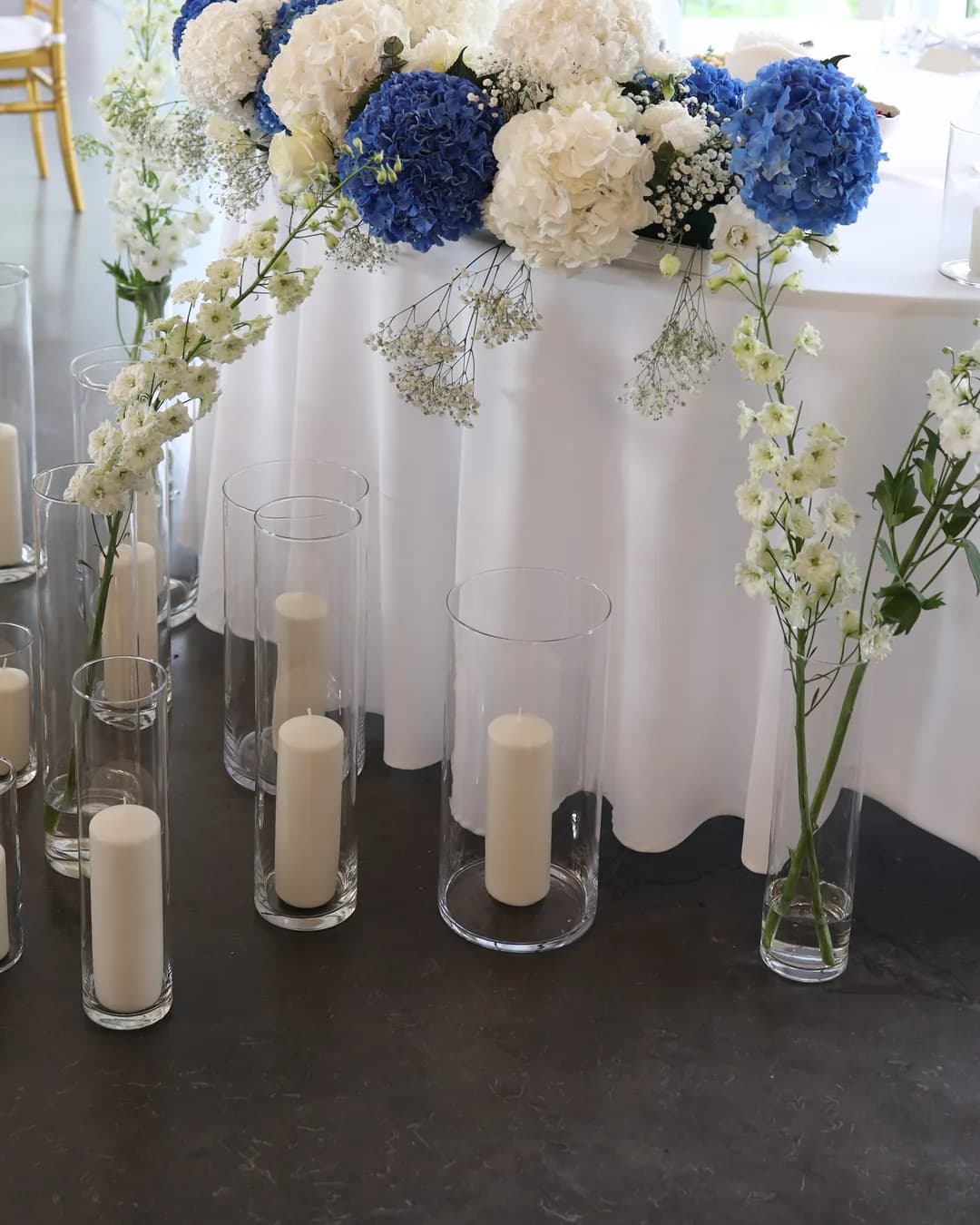 Pillar candles in glass cylinders of different heights and vases with white delphinium in front of the bride and groom's table