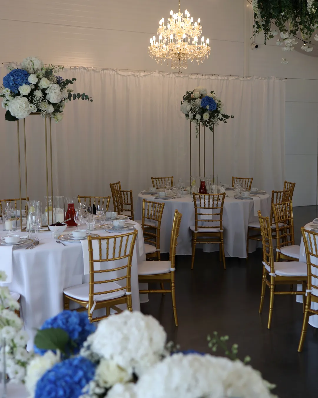 A room with round tables and tall compositions of hydrangeas on golden flower stands, a low composition in the foreground