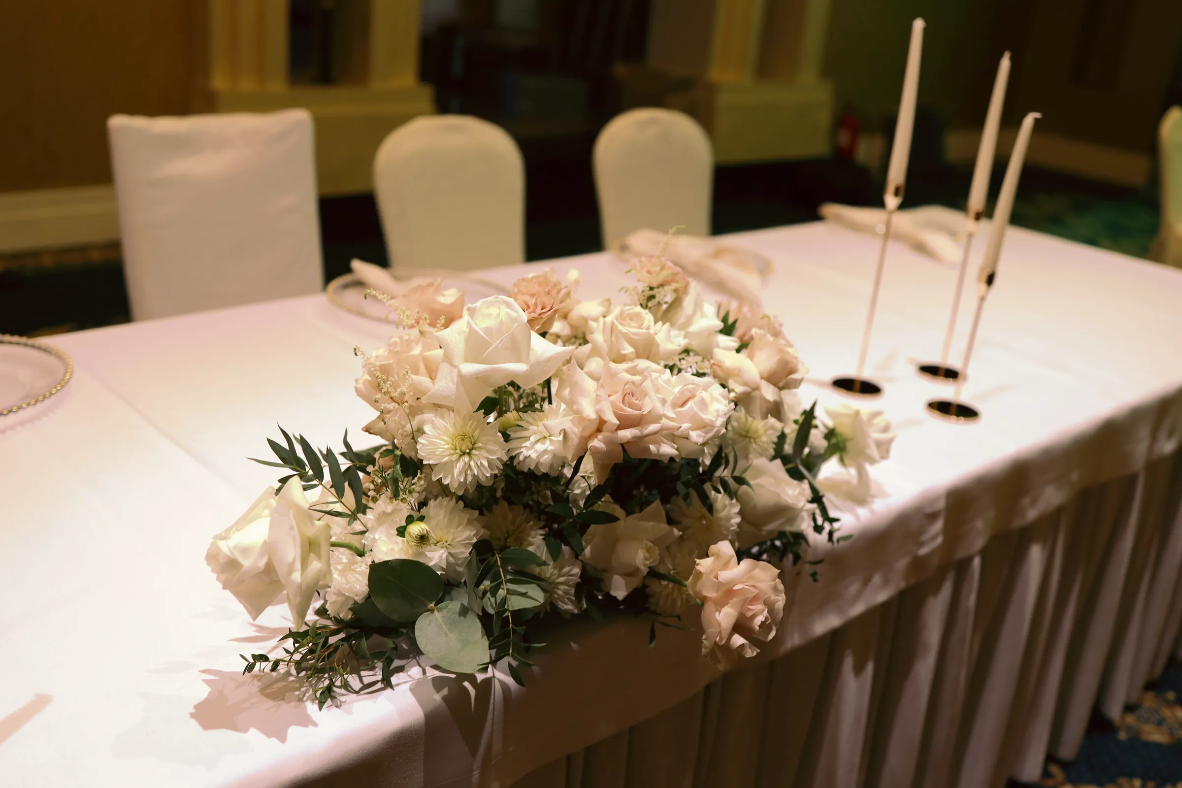 A composition on the bride and groom's table with cream and powder roses, white chrysanthemums and eucalyptus next to three tall candles