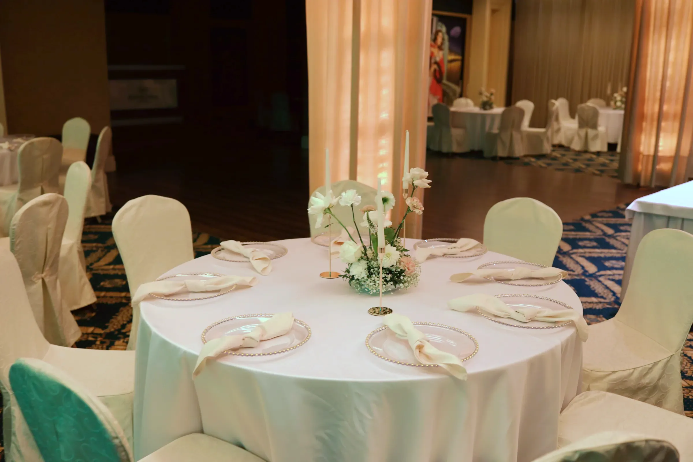 A round table with a white composition, candles and napkins on glass charger plates, other tables and columns of the hall in the background