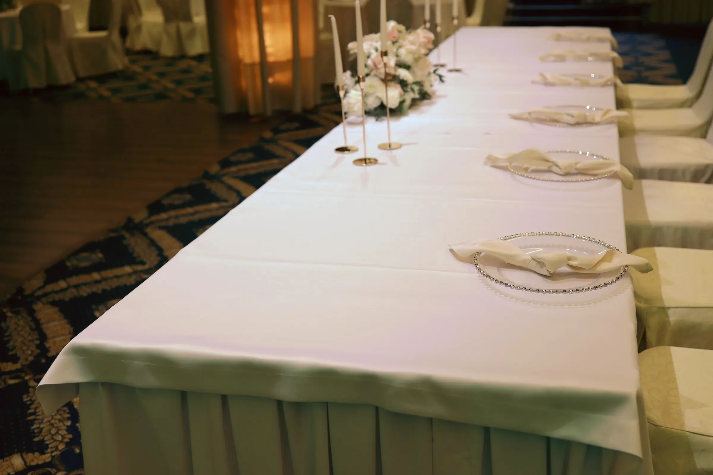 Long table of the bride and groom with a white tablecloth, glass plates, cream napkins and candles next to a rose arrangement