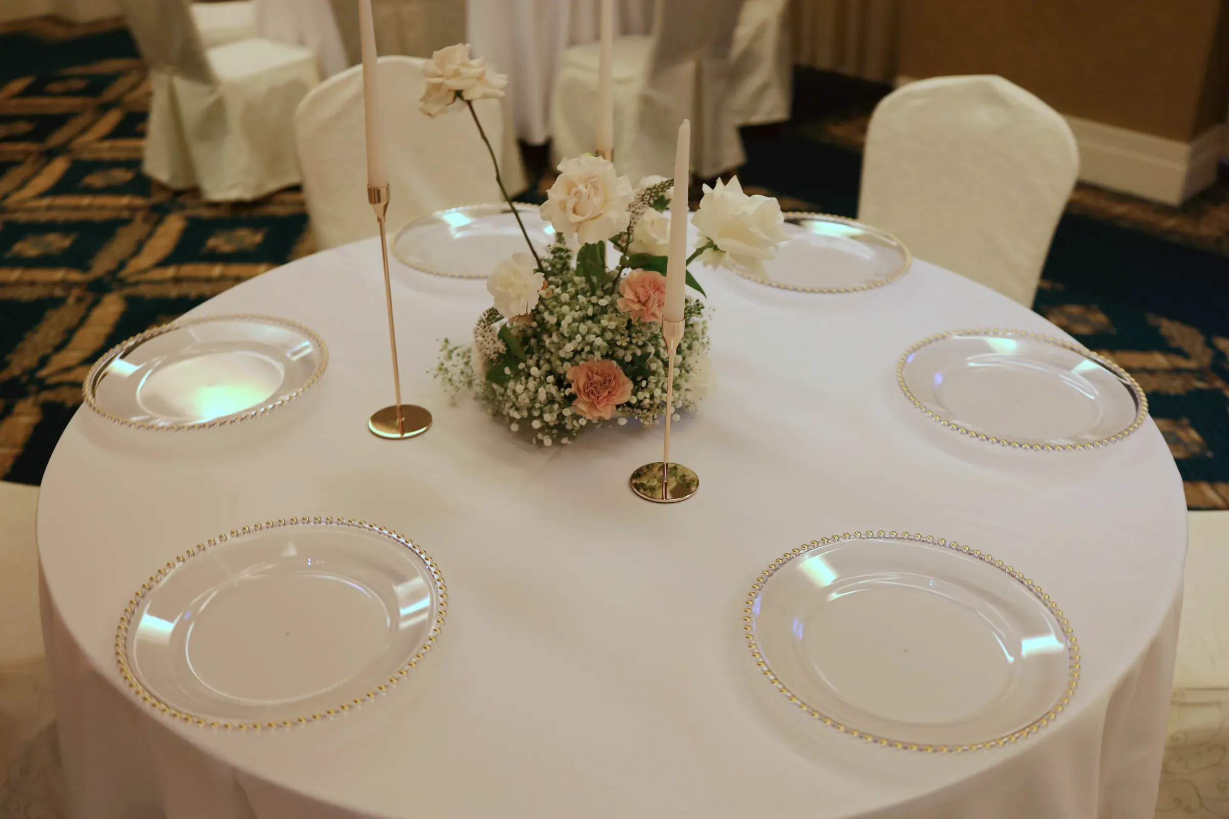 Round table with glass plates with a gold edge, candles in gold candlesticks and a composition with peach carnations
