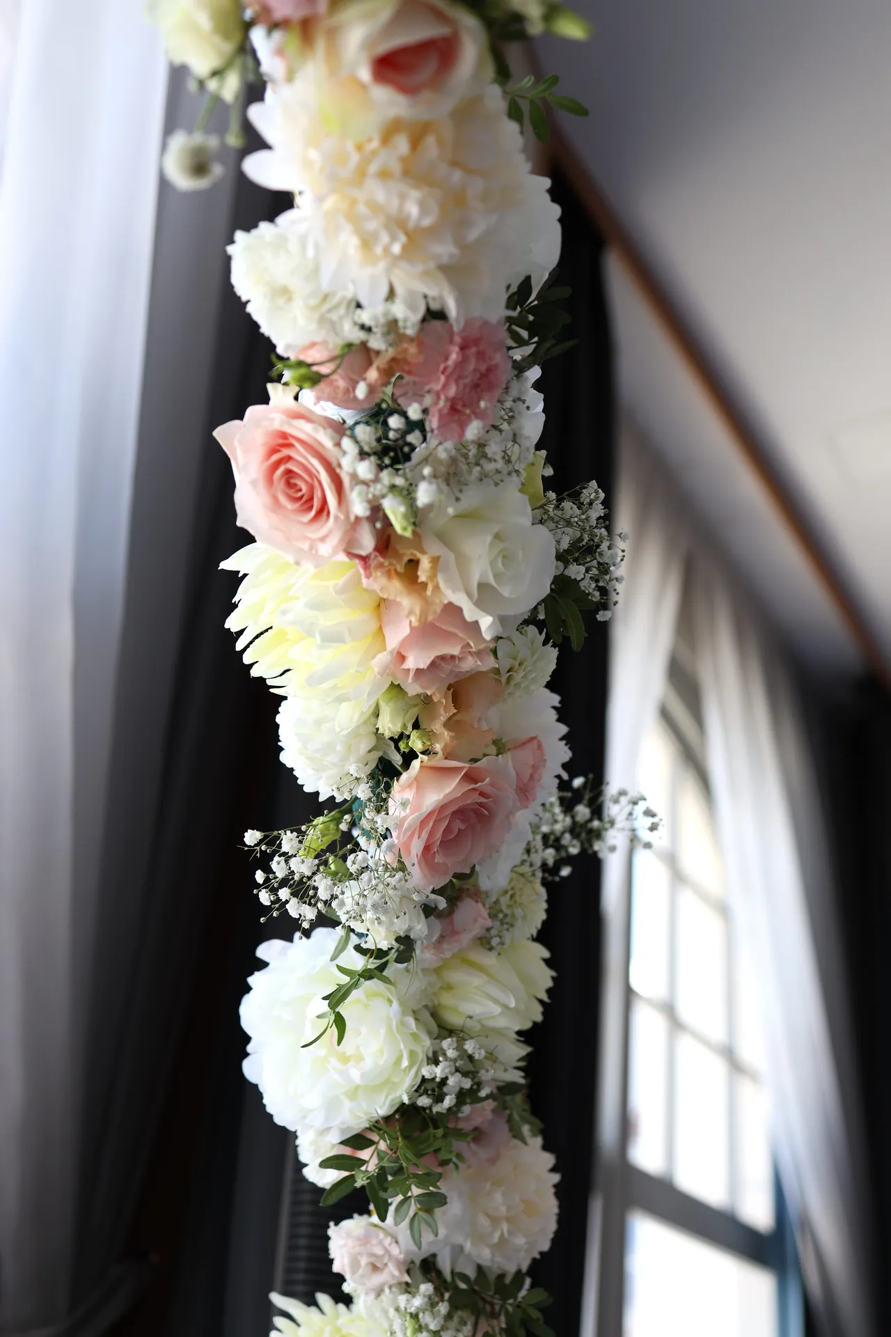 A strip of white and pink roses with greenery on the rim that runs along the arched window and dark curtain
