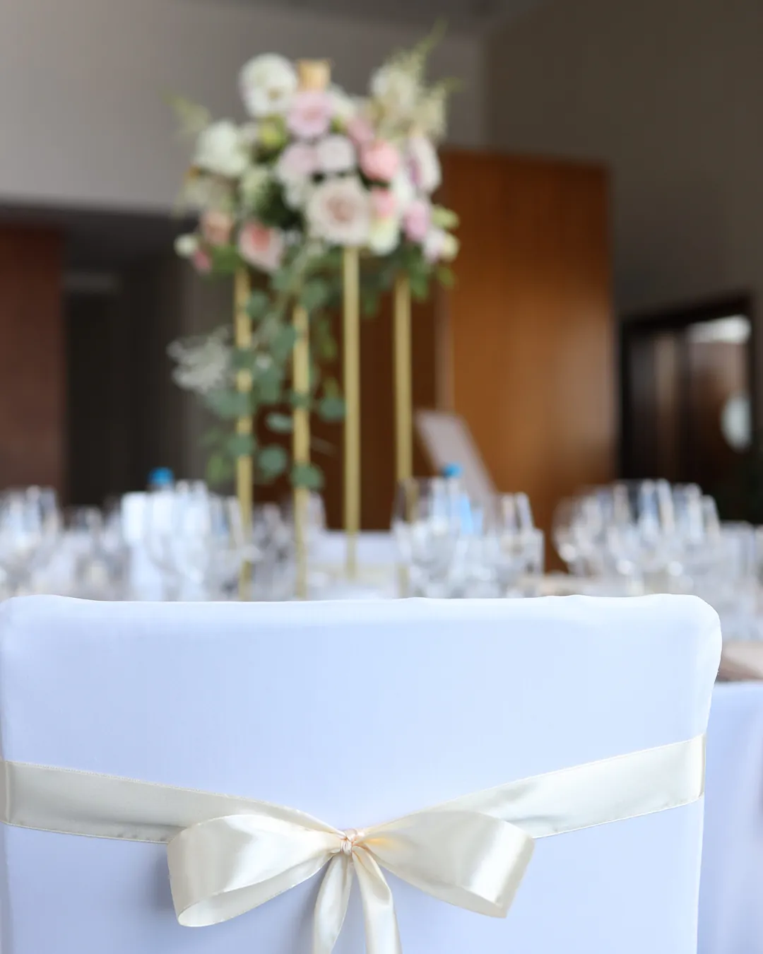 A chair in a white cover with a cream bow, and behind it a golden stand with flowers on a set table