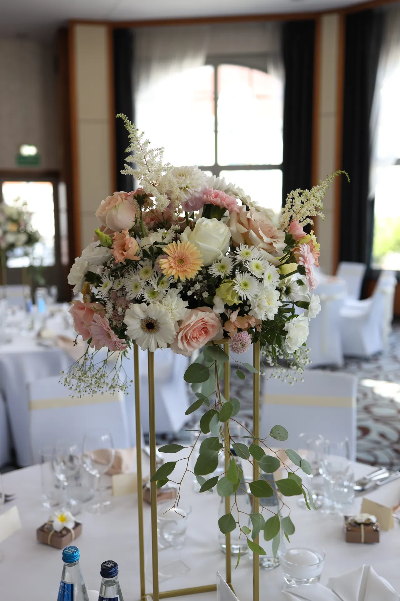 A bouquet in white and powder pink on a slim gold stand placed on a round table near the windows