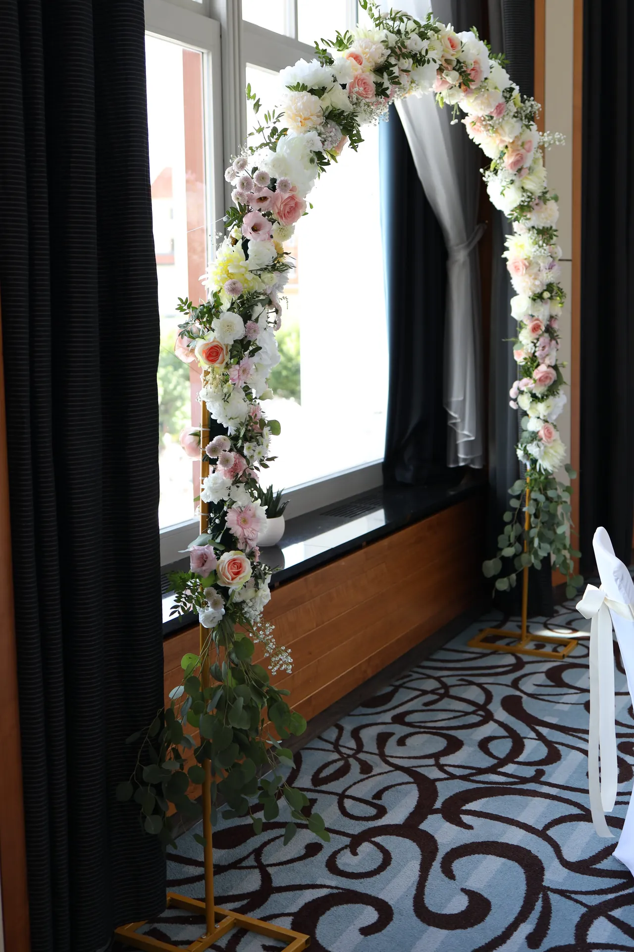A hoop of white and pink flowers with greenery stands next to an arched window and frames the view beyond the glass