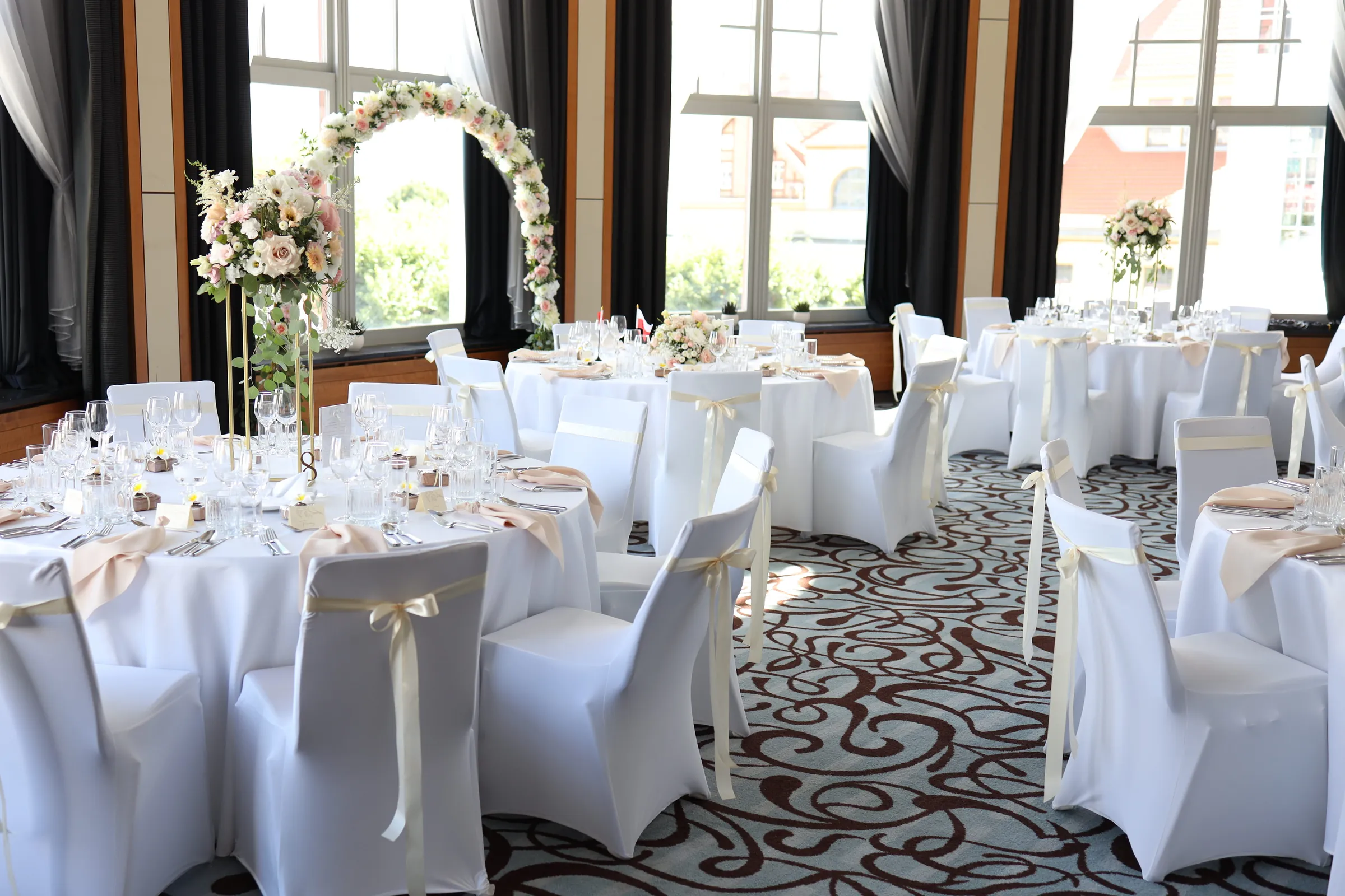 Round tables in white, chairs with cream bows and a flower hoop by the arched window of the room