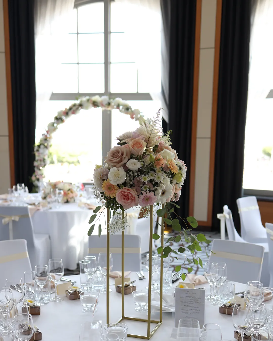 A tall gold stand with white and pink flowers on the table, and behind it a flower hoop by the arched window