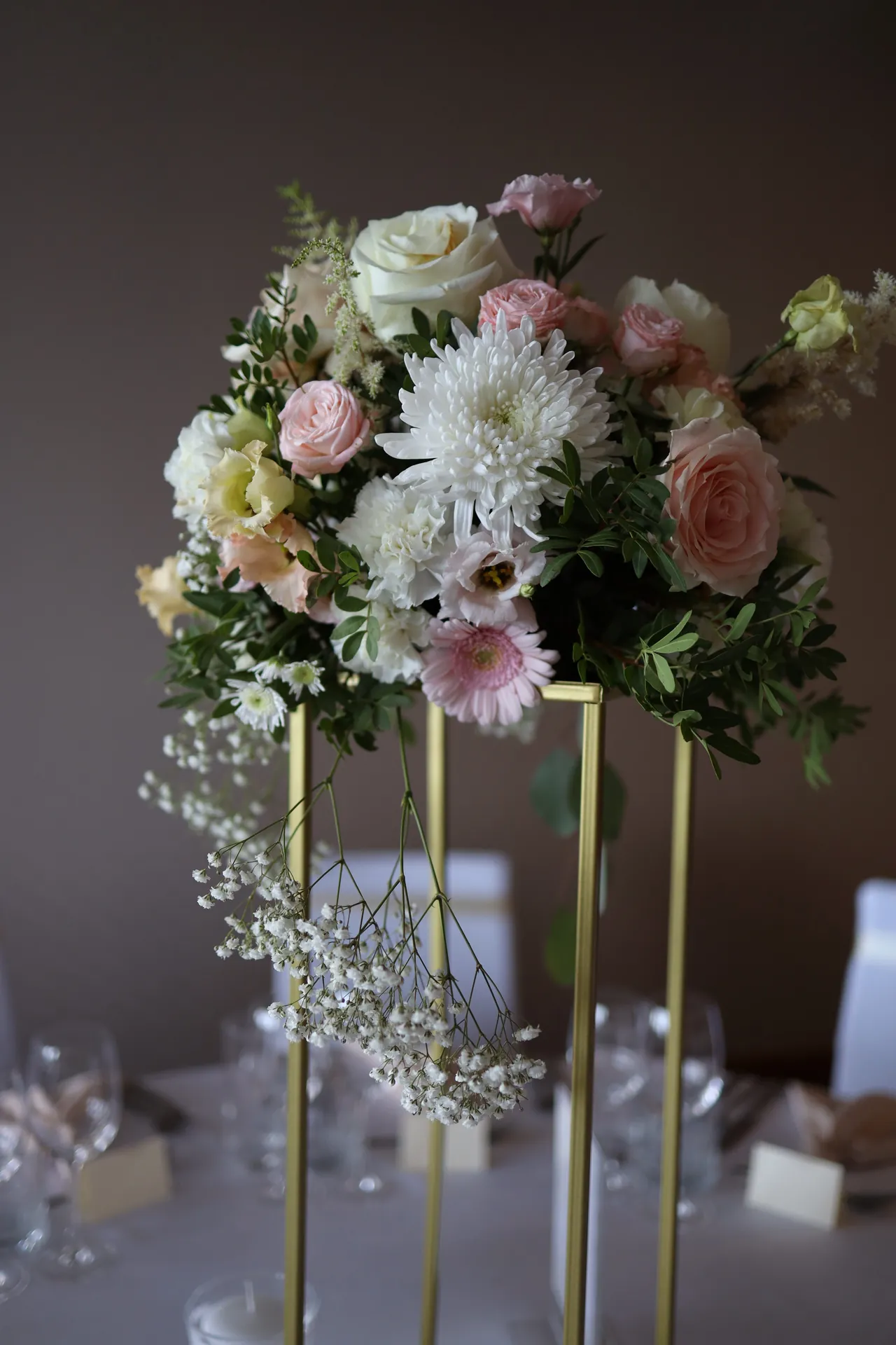 Blush roses, white flowers and gypsophila on a tall golden stand against the dark wall of the room