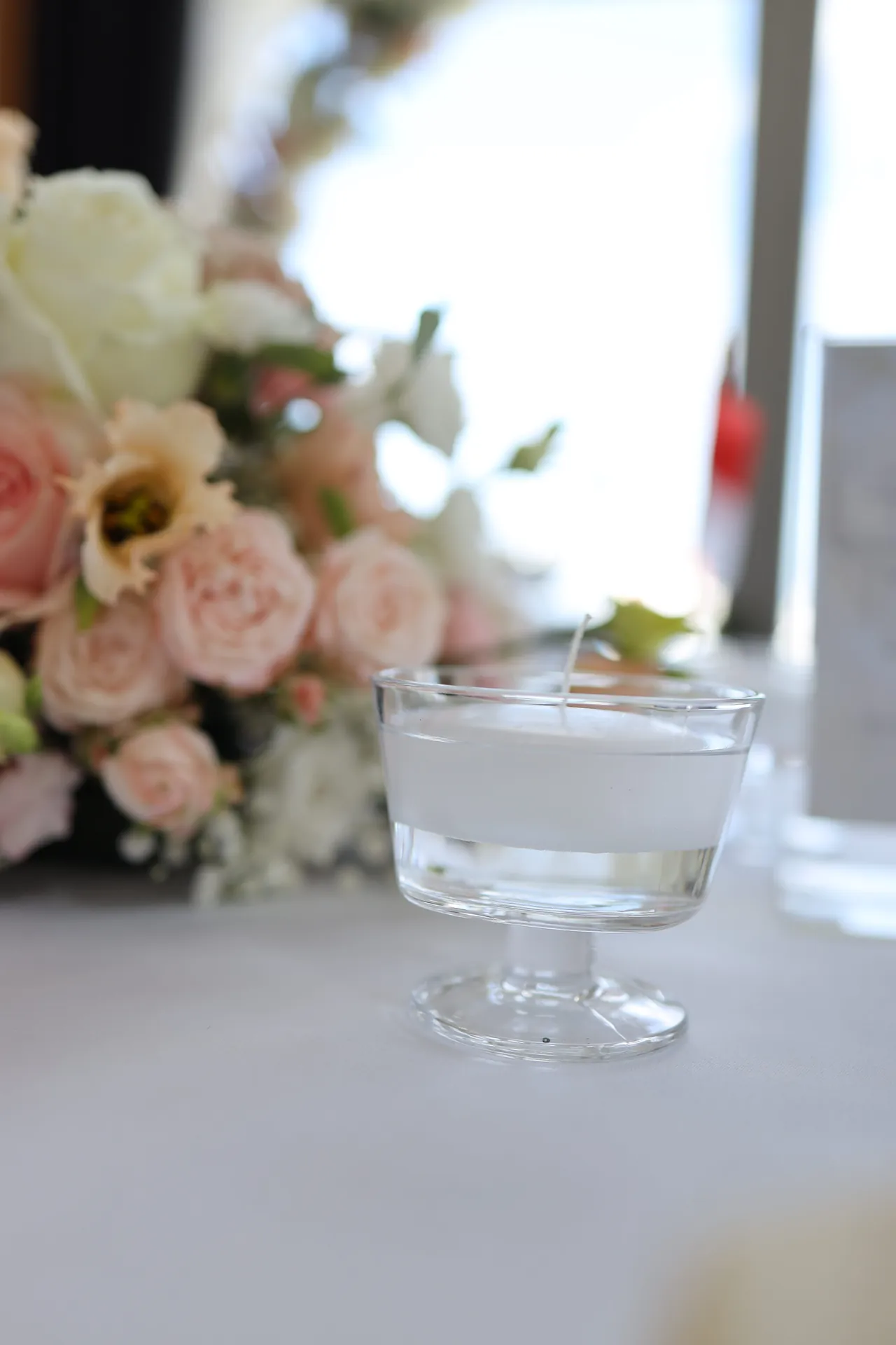 A candle in a glass cup on a white tablecloth next to a composition of blush roses and white flowers