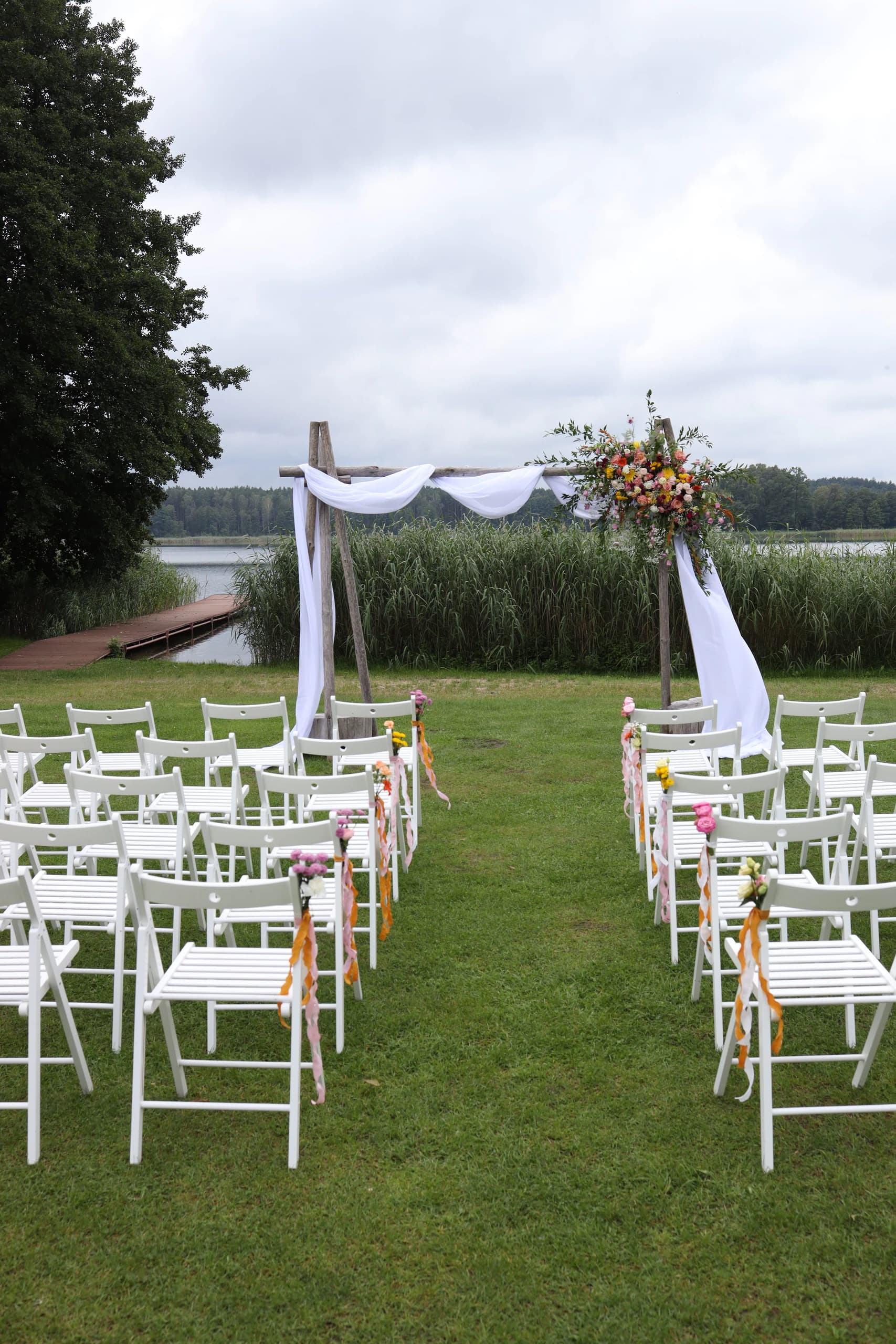 The passage between the white chairs leads to an arch, on the outermost chairs there are bouquets of long ribbons, on the left there is a pier and a lake