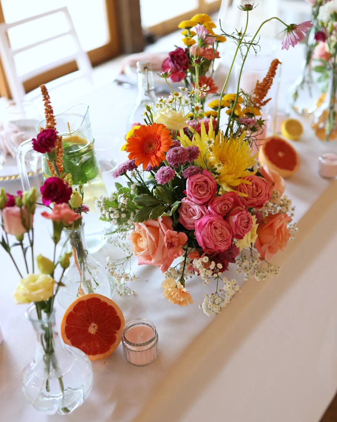 A lush composition on a rectangular table: gerbera, roses, spider chrysanthemum and carnations, in front of it a grapefruit, a candle and a vase