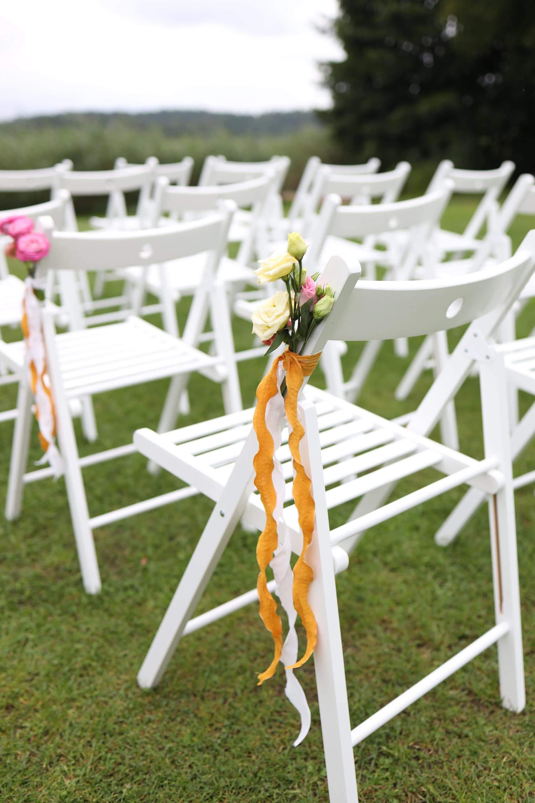 White chair with a bouquet of cream eustomas and a pink bud, mustard orange and white wavy ribbon fall into the grass
