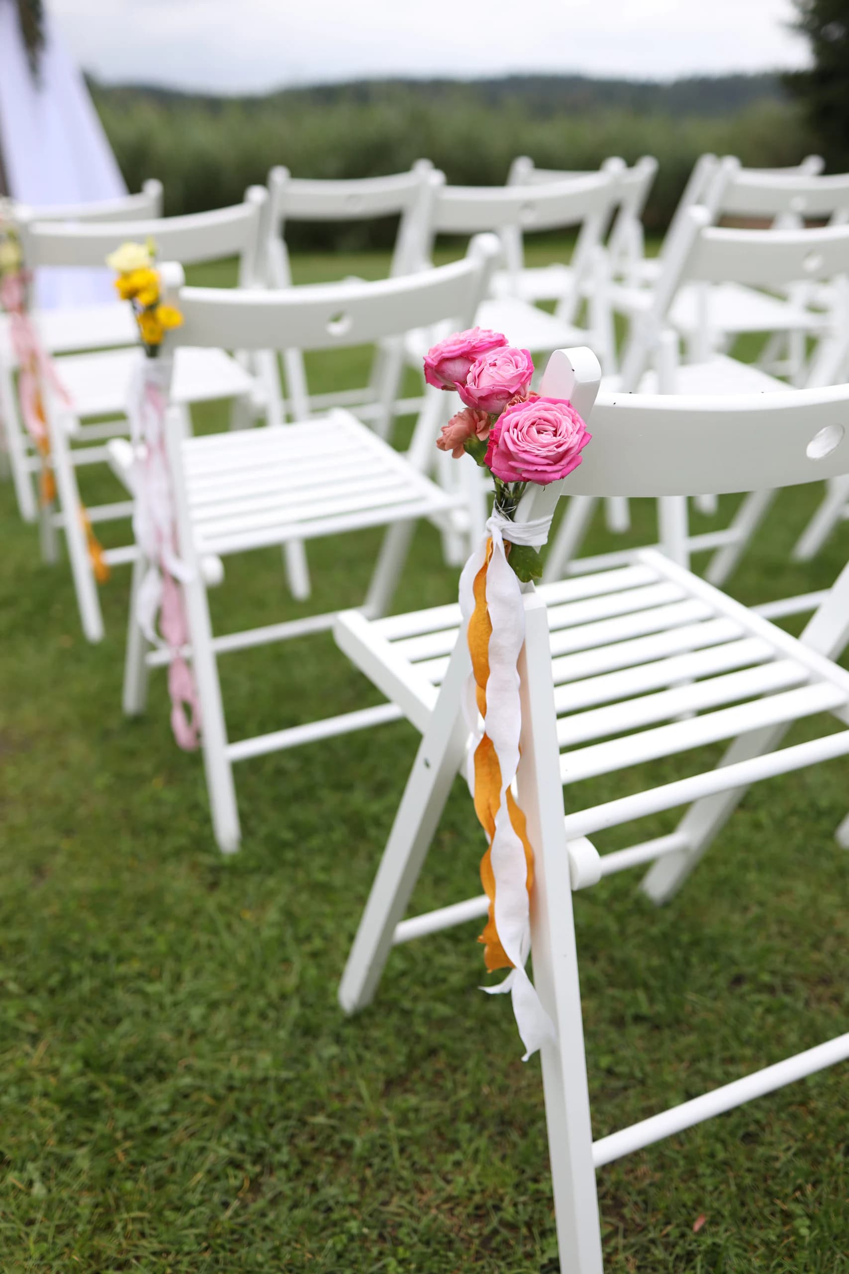 White chair: a bunch of pink spray roses, a white and mustard orange ribbon, a chair with yellow flowers and a pink ribbon in the background