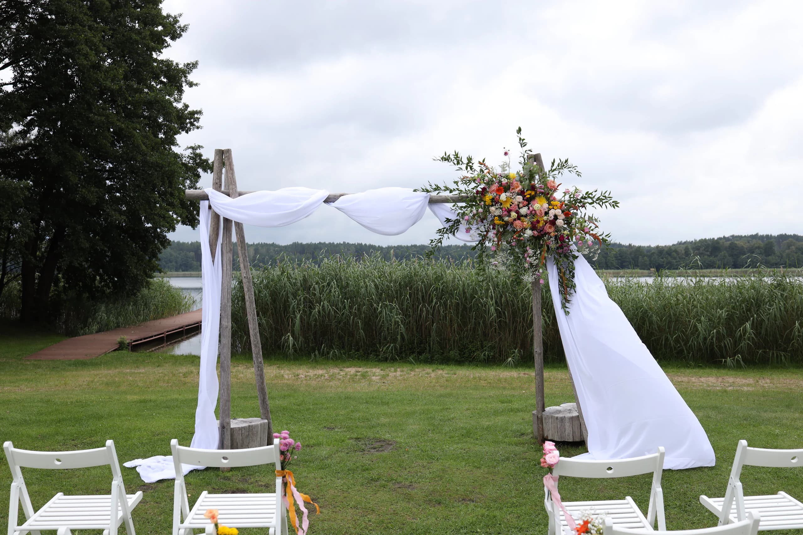 Wedding arch with poles on stumps: white fabric in two waves and flowers in the upper right corner, behind the arch there are reeds, a pier and a lake