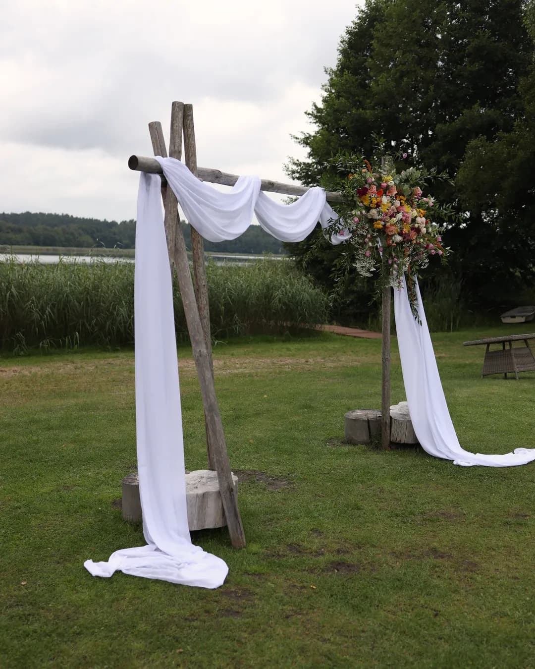 A wedding arch made of poles with white fabric and an arrangement in the upper right corner, legs in stumps, behind it reeds, a lake and a large tree