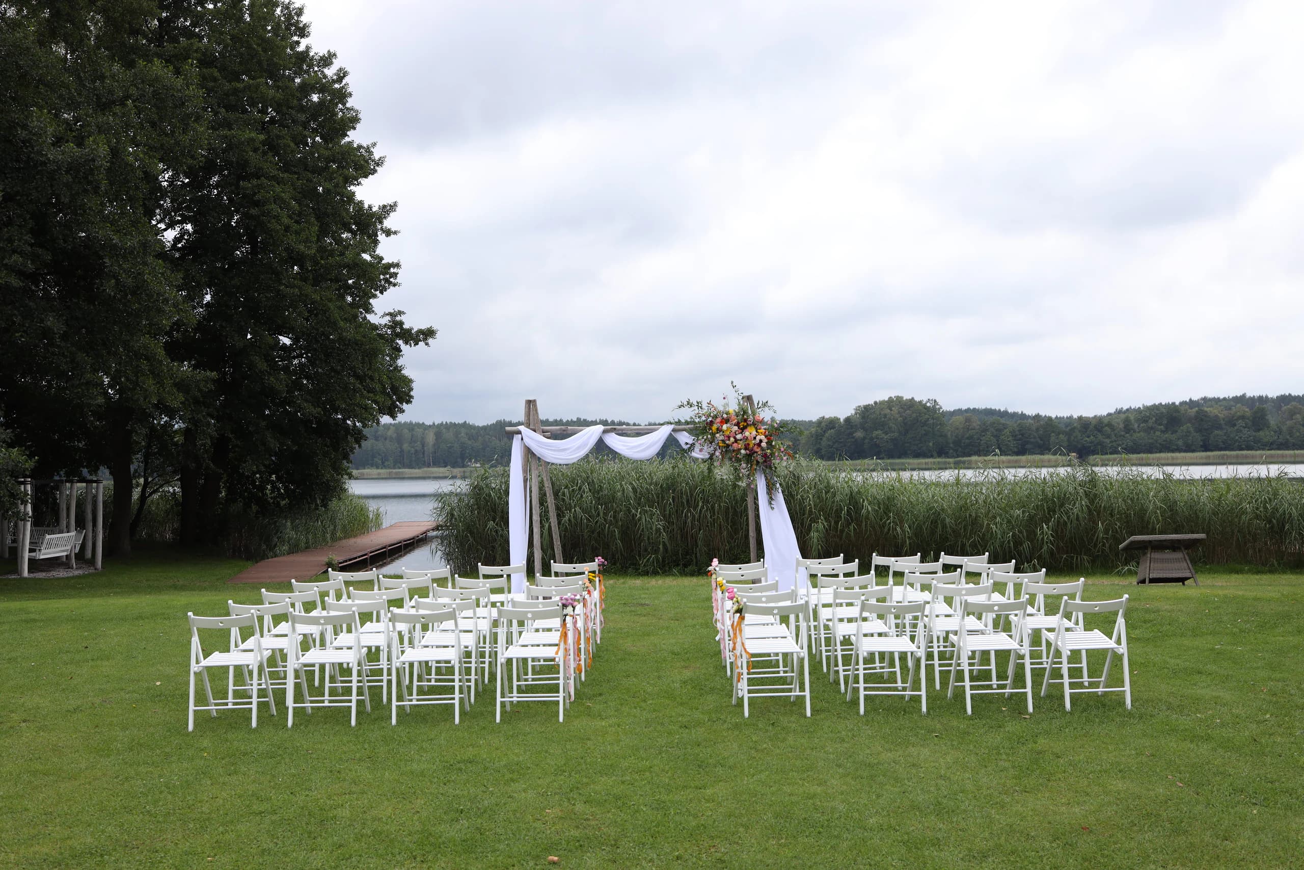Outdoor wedding by the lake: arch with fabric and flowers, two blocks of white chairs with an aisle, a large tree on the left, reeds and a forest in the background