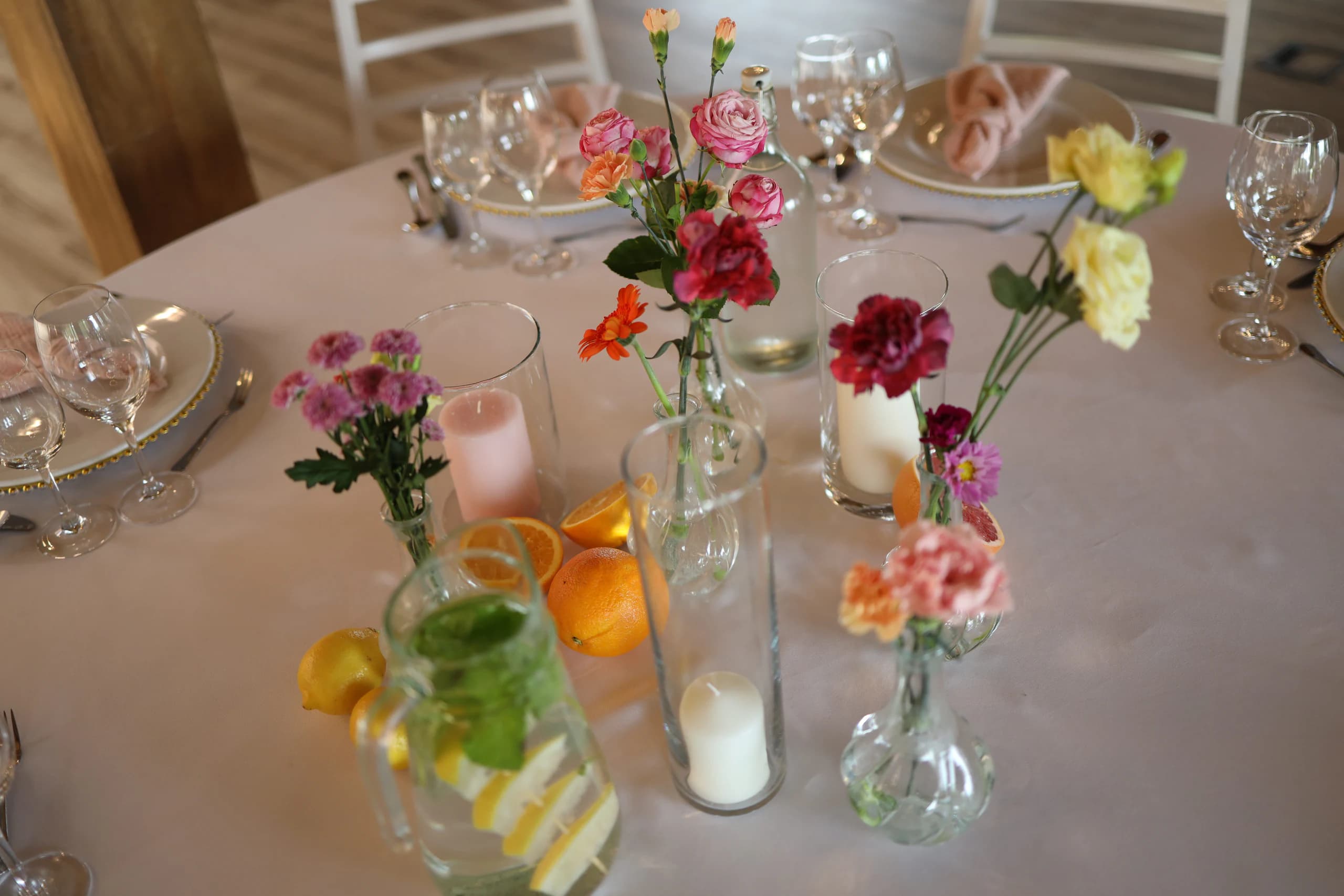 Table from above: pink and white candles in glass cylinders, vases with spray roses, carnation and yellow eustoma, jug and citrus fruits