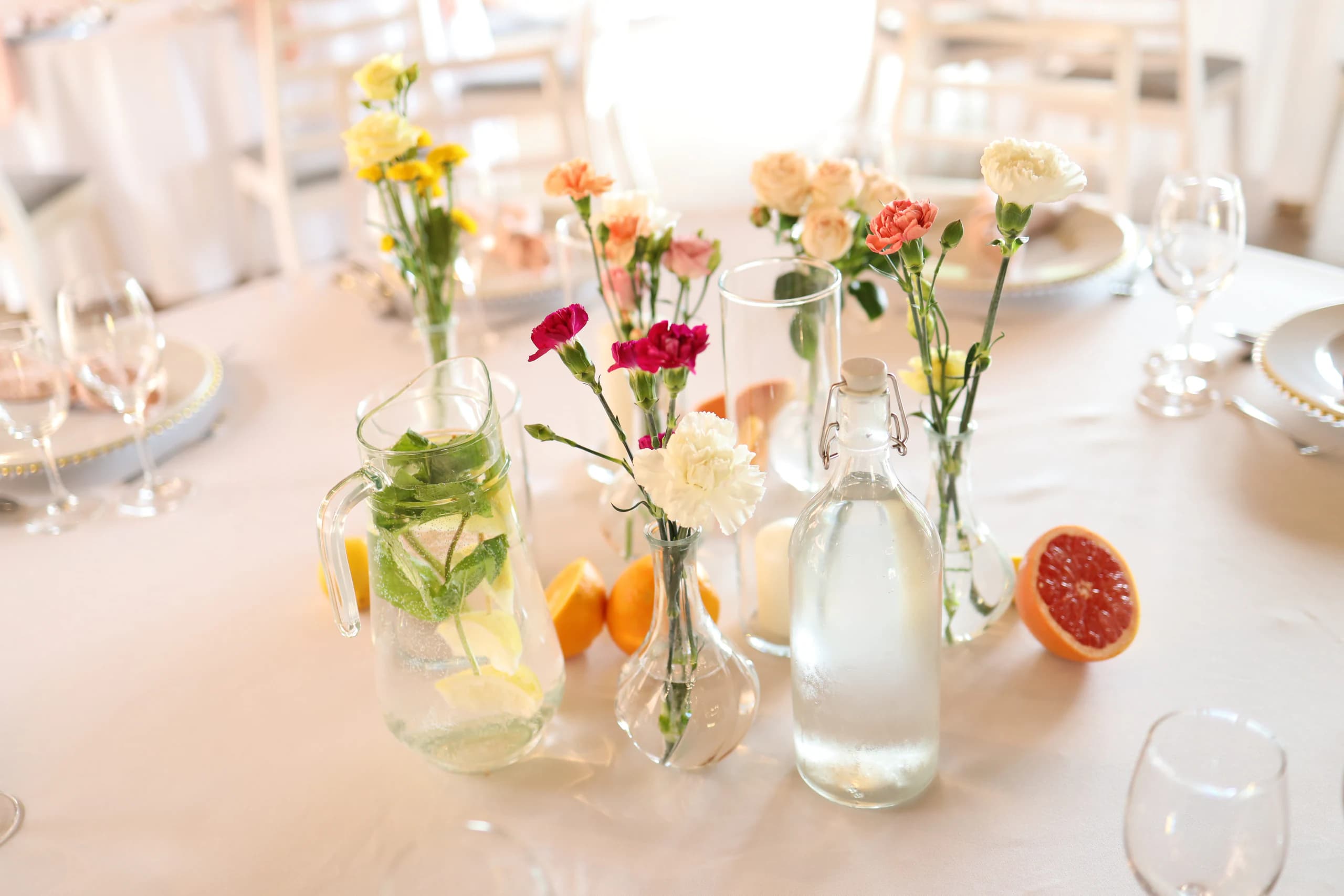 Vases with single carnations, spray roses and yellow flowers, jug with lemon and mint, half of a grapefruit and oranges