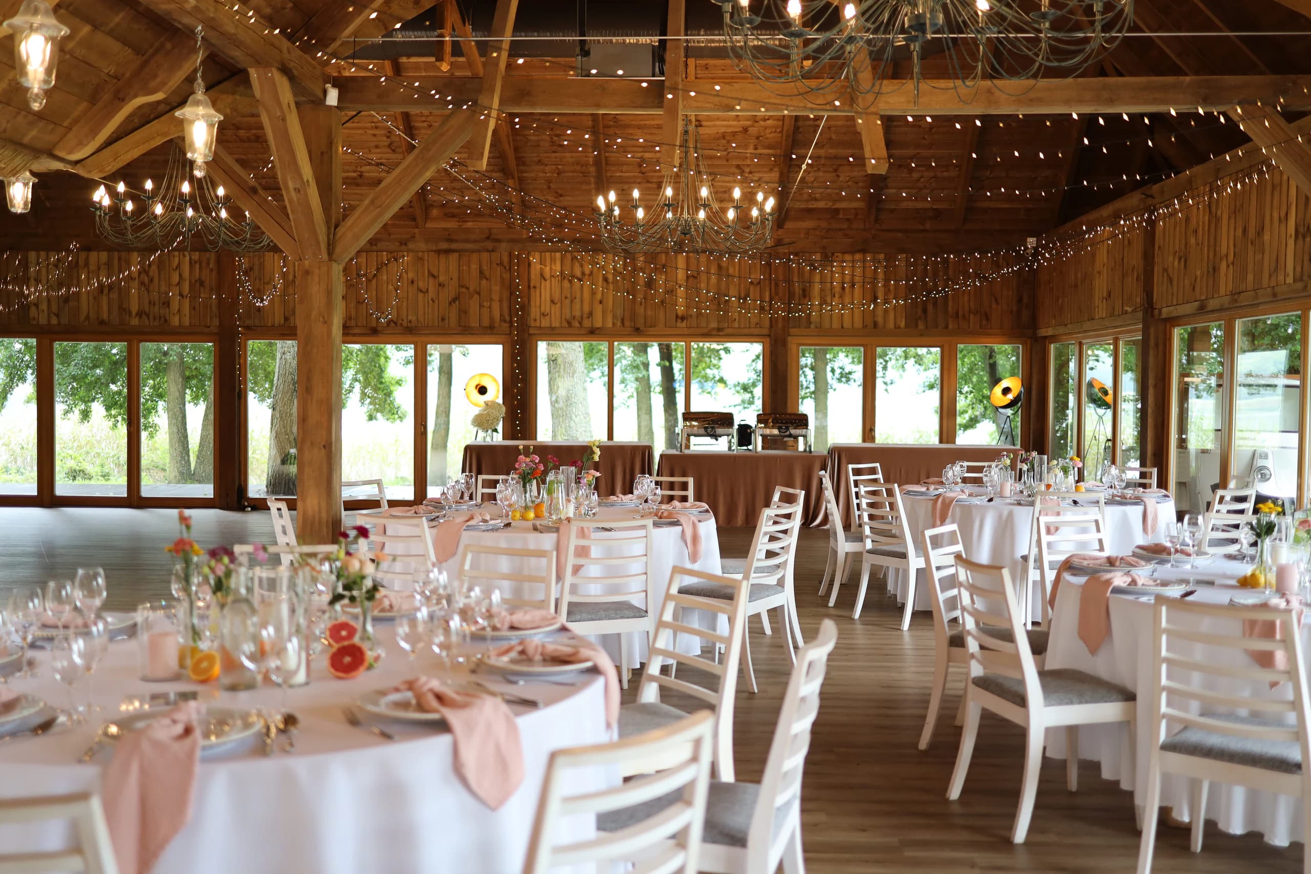 A wooden room with an exposed truss, chandeliers and lamps, glass walls, round tables with white tablecloths and vases with flowers.