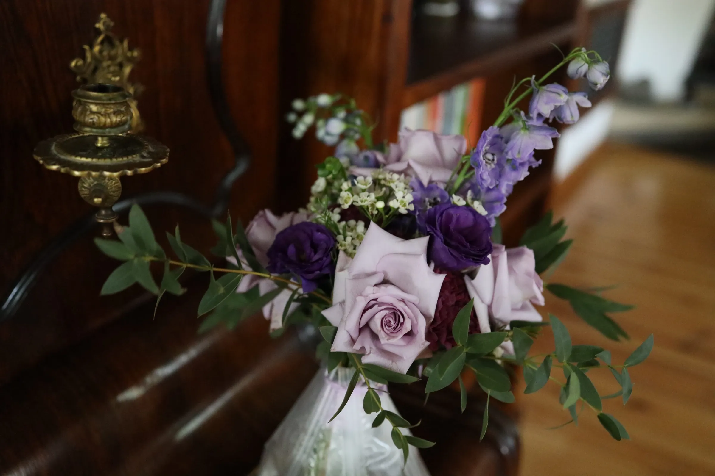 A bouquet of lilac roses, purple eustoma and delphiniums in a vase on a piano, next to a brass candlestick