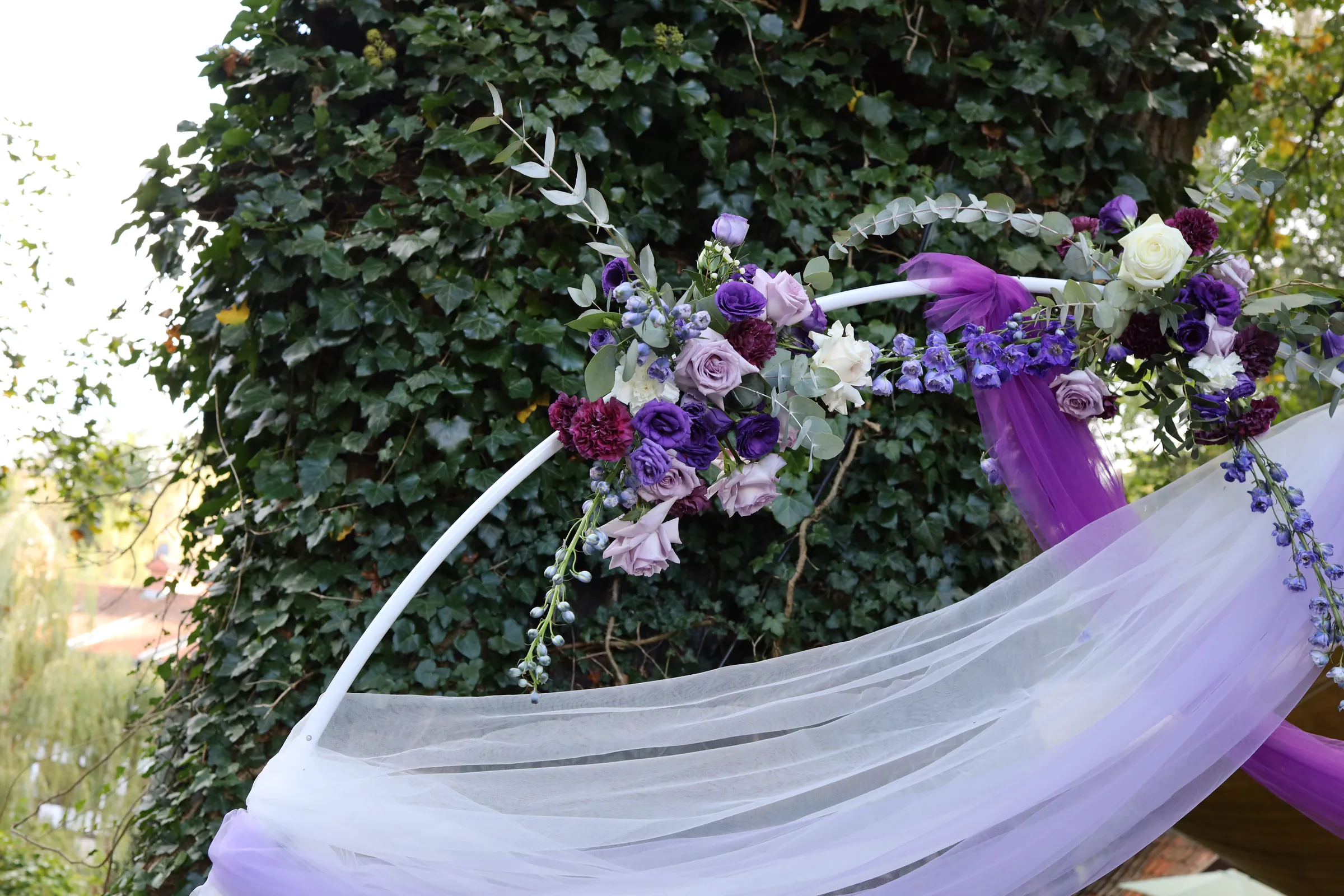 Roses, eustoma, carnations and delphiniums with eucalyptus on the hoop arch, under them white and lilac tulle on a background of ivy